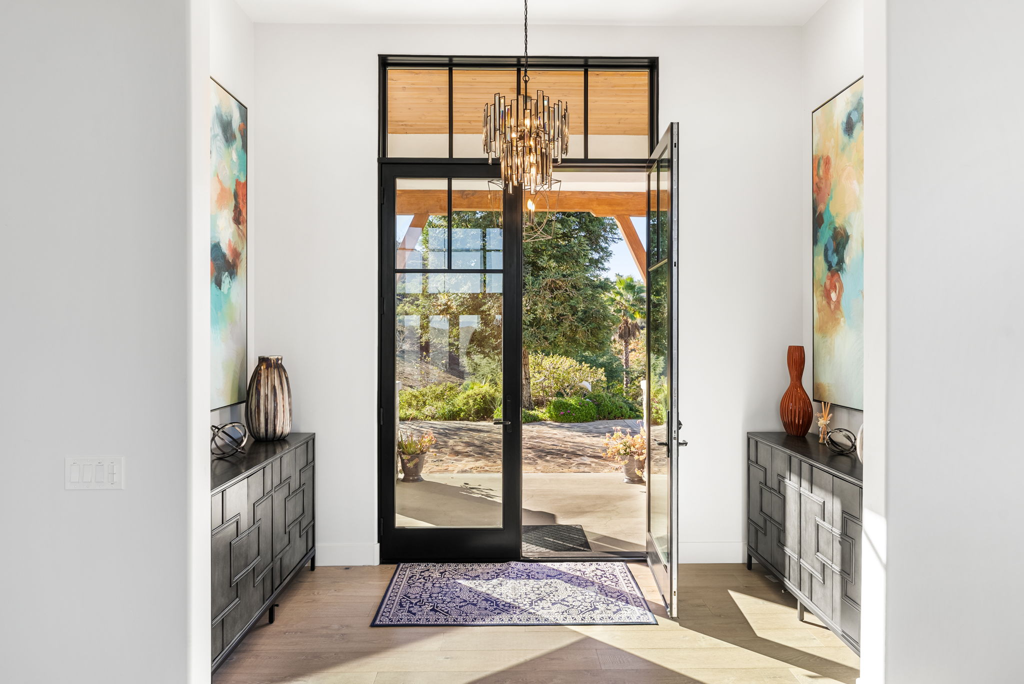 This elegant entryway features a symmetrical layout with matching dark-toned cabinets, abstract artwork, and decorative vases on either side of a grand, floor-to-ceiling iron and glass front door. A sophisticated multi-tiered chandelier serves as a focal point, while the light hardwood flooring and neutral color palette create a warm, inviting, and modern atmosphere. The open door reveals a picturesque view of the outdoor patio and landscaped yard, seamlessly connecting the indoor and outdoor living spaces.
