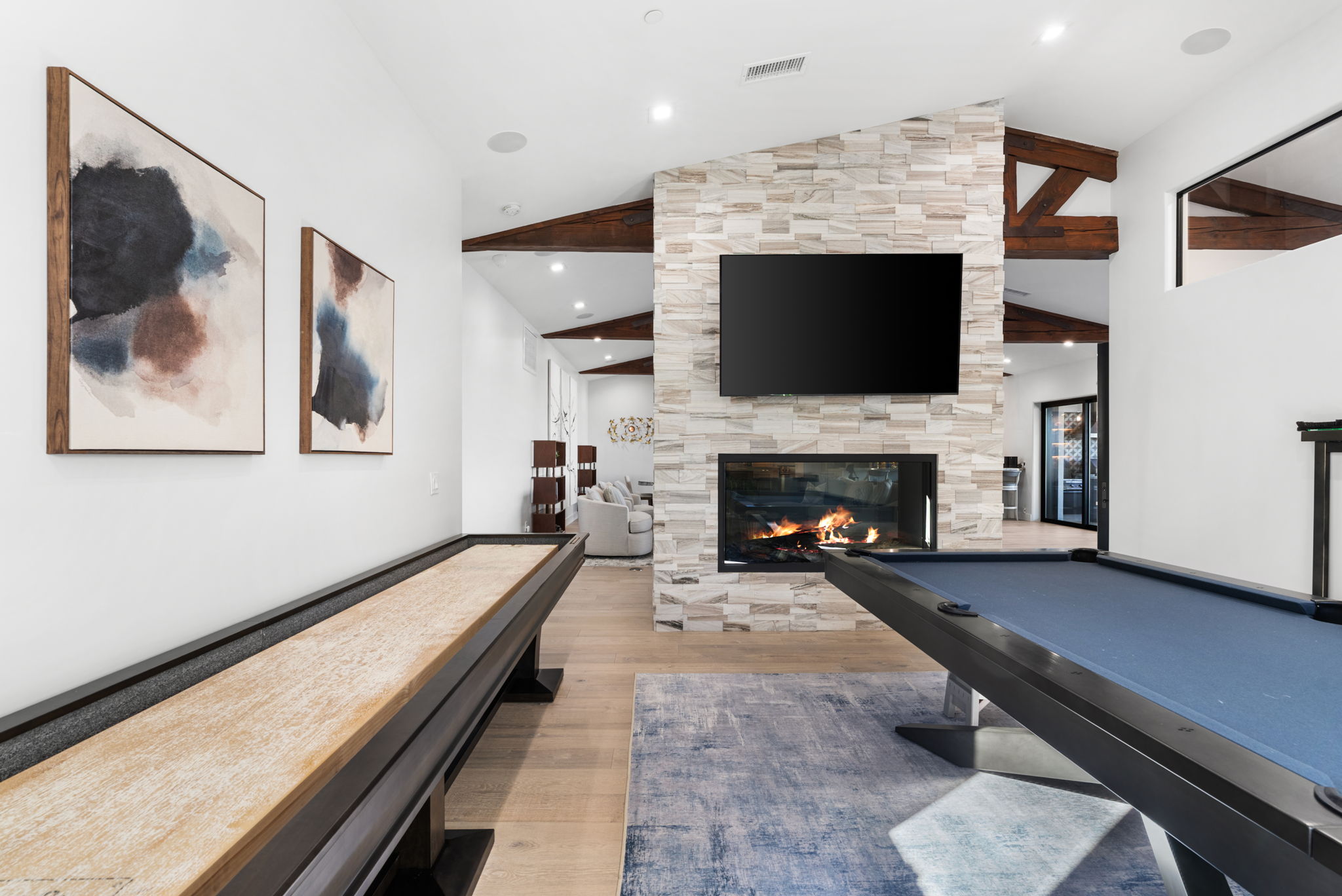 This interior space serves as a dedicated game room, featuring a professional-grade pool table on a blue rug and a lengthy shuffleboard table. A central stone fireplace with an integrated television adds a cozy focal point, while exposed wooden beams and artwork provide a sophisticated, recreational aesthetic. The open floor plan creates a seamless transition between the gaming area and the adjacent living space.