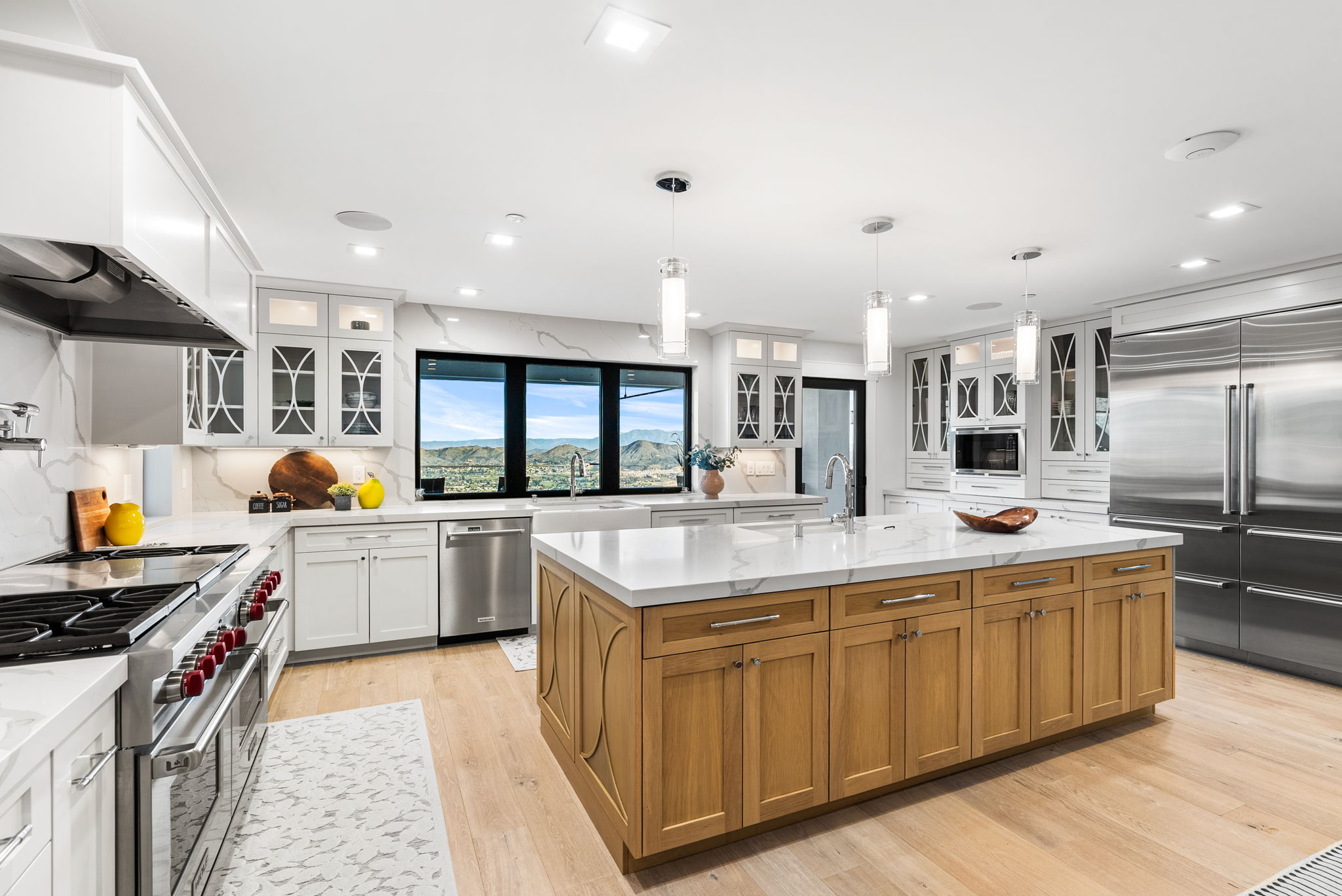 This stunning gourmet kitchen features a harmonious blend of white upper cabinetry and a warm, natural wood-toned center island with quartz countertops. The space is equipped with professional-grade stainless steel appliances, including a large refrigerator and a heavy-duty range, all complemented by modern pendant lighting. Large windows frame a scenic view of the surrounding mountains, providing both natural light and an impressive focal point for the open, contemporary layout.