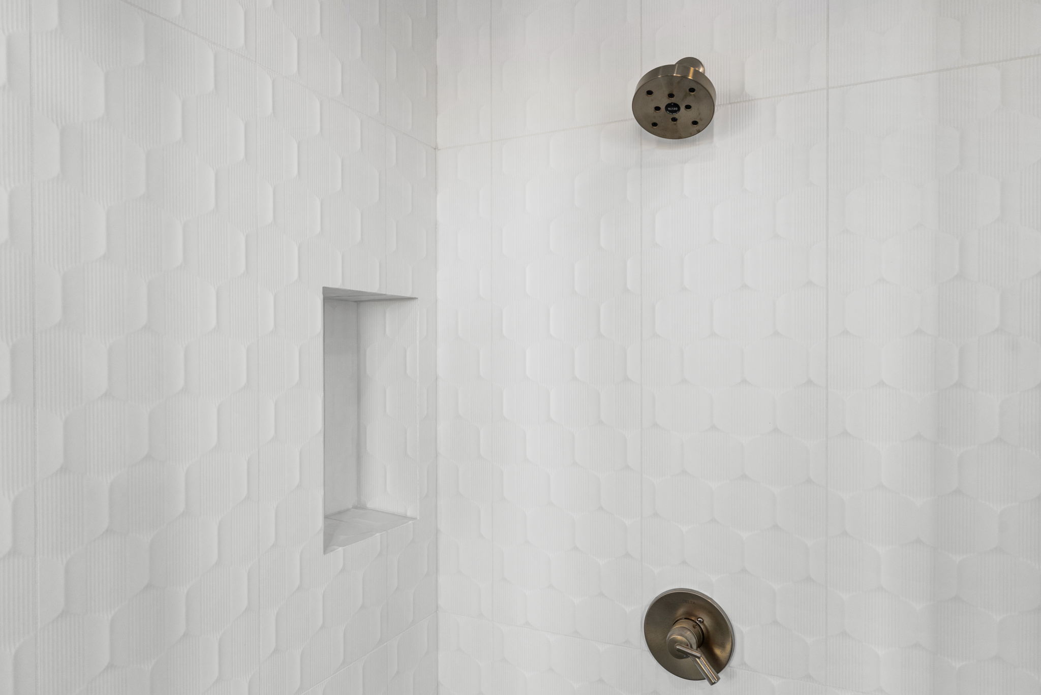This shower area features a contemporary design with textured, hexagonal white wall tiles that add subtle visual interest. It includes a built-in recessed wall niche for storage and high-quality, brushed-nickel shower fixtures, creating a clean, spa-like aesthetic.