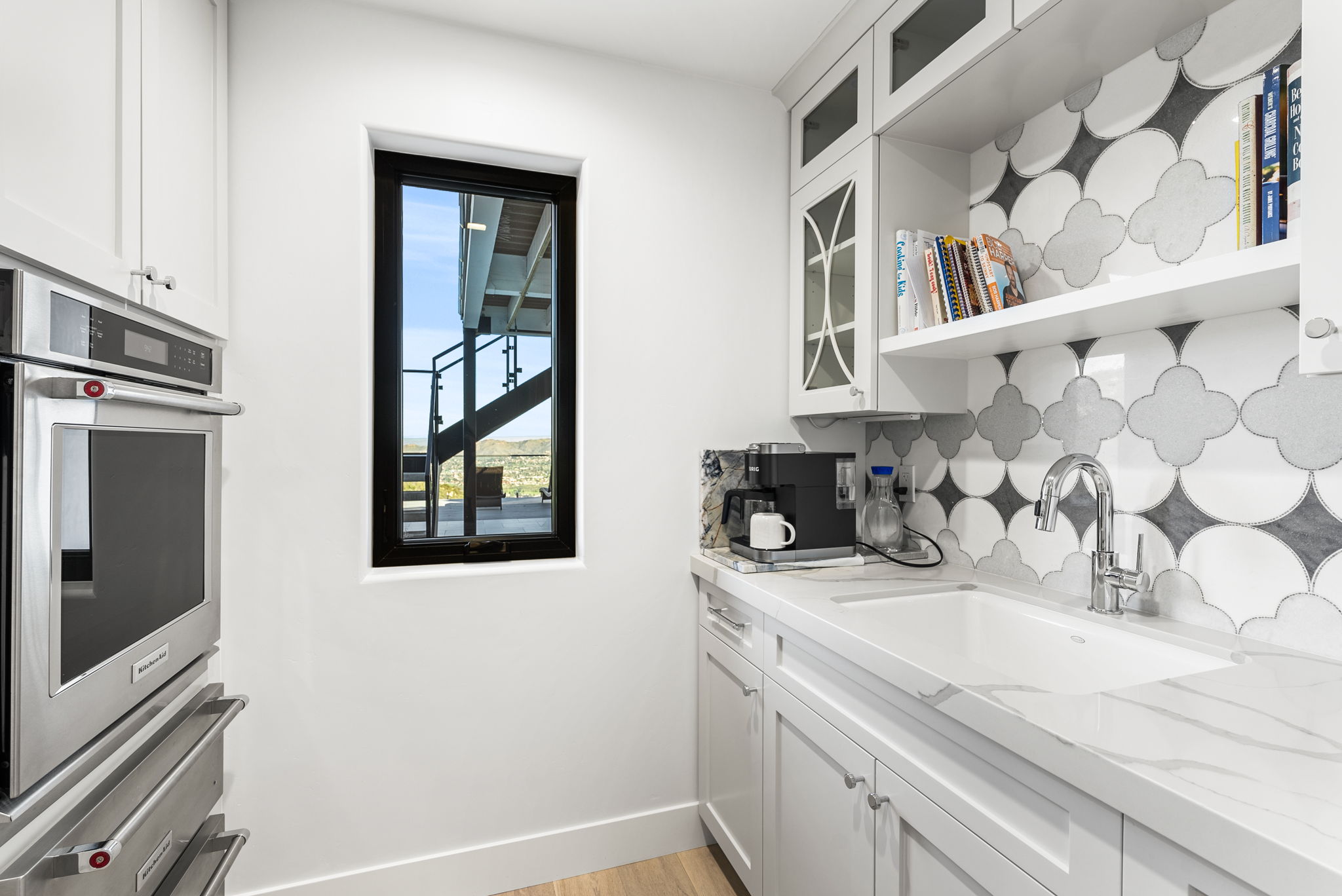 This bright and functional wet bar or butler's pantry area features crisp white cabinetry, marble-style countertops, and a striking grey and white patterned backsplash. A sleek undermount sink, a built-in coffee station, and a tall KitchenAid wall oven define the space, while a narrow window provides a view of an outdoor deck and staircase. The clean, modern design creates a sophisticated transition space that is both practical and visually appealing.