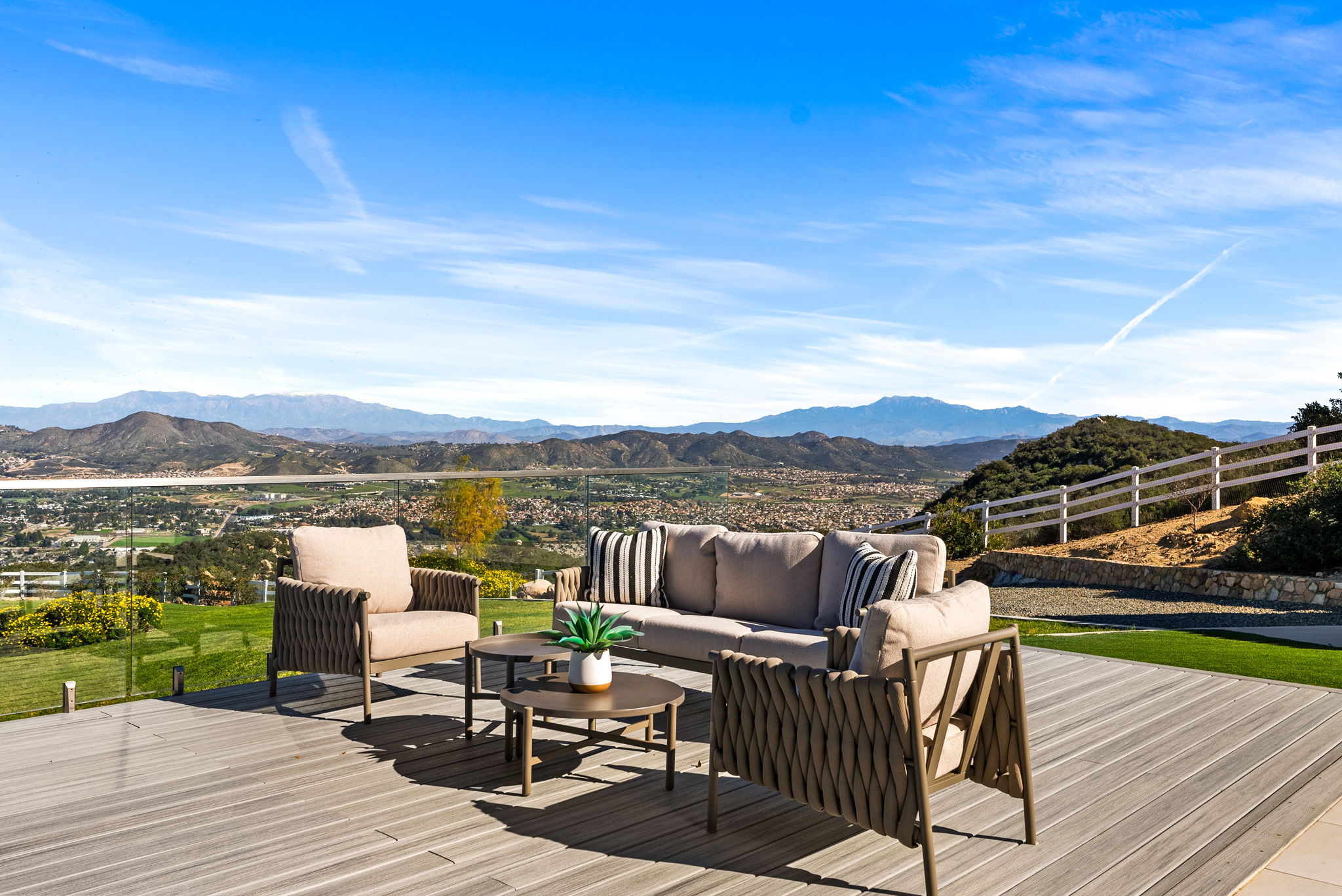 This stunning outdoor deck features modern lounge furniture, including a plush sofa and matching armchairs, arranged to maximize the breathtaking panoramic view of the valley and distant mountain ranges. The sleek, glass-railing design ensures an unobstructed perspective, creating an ideal space for relaxation and entertaining. The sophisticated, contemporary aesthetic is complemented by a manicured lawn and a clear blue sky, making it an exceptional highlight of the property.