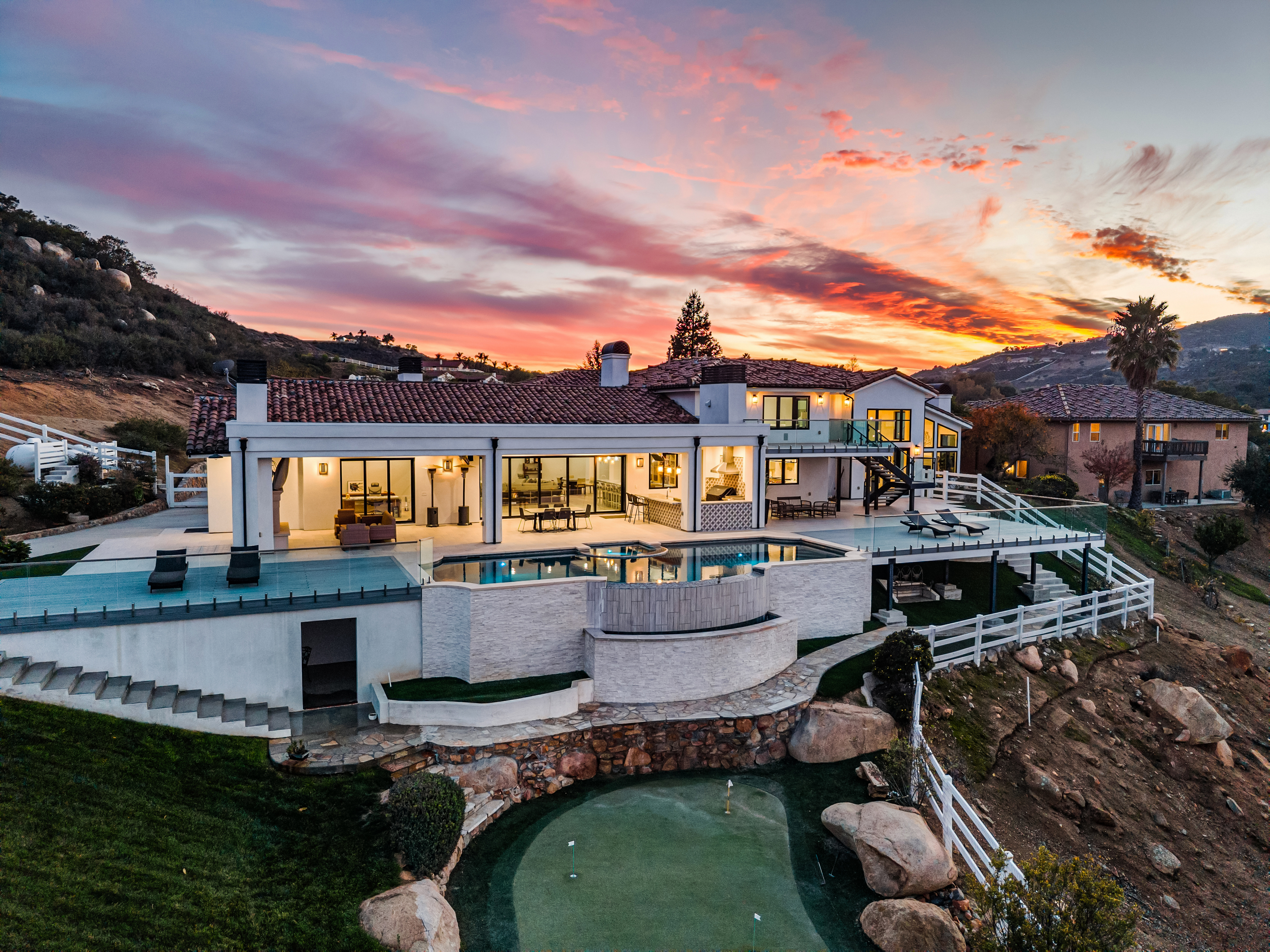 This stunning luxury estate is captured from an aerial perspective, showcasing a sprawling multi-level backyard designed for high-end outdoor living. Key features include a seamless infinity-edge pool, an expansive composite deck with glass railings, and a professional-grade putting green set against a picturesque sunset. The home exhibits a contemporary architectural style with a traditional tiled roof, perfectly blending indoor-outdoor flow.