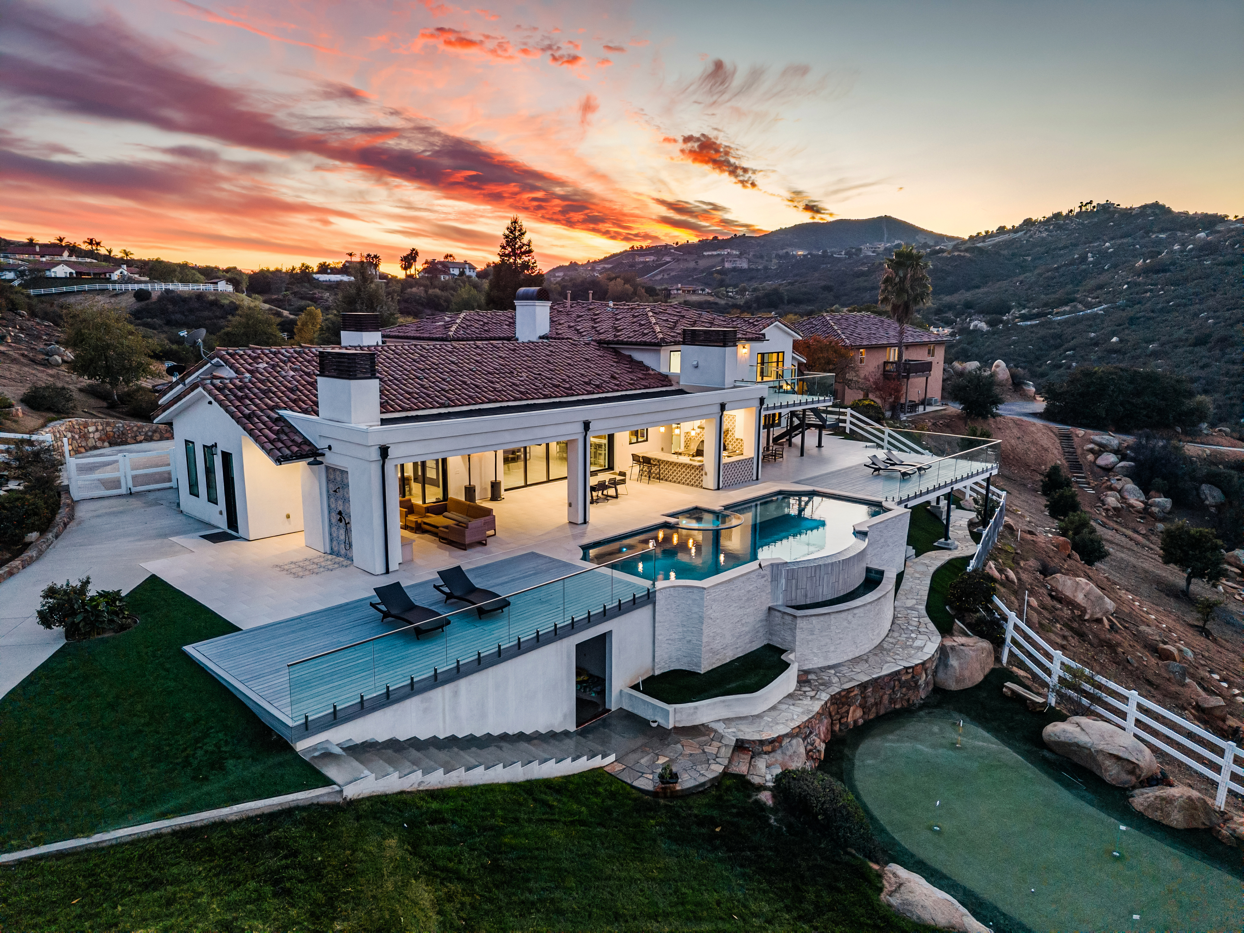 This breathtaking aerial view showcases a luxury estate property during a vibrant sunset. The home features a seamless indoor-outdoor living design with a sprawling terrace, an infinity-edge pool, and a private putting green nestled against a rugged hillside. The architectural style blends modern refinement with classic Mediterranean elements, offering expansive views and premier outdoor amenities.
