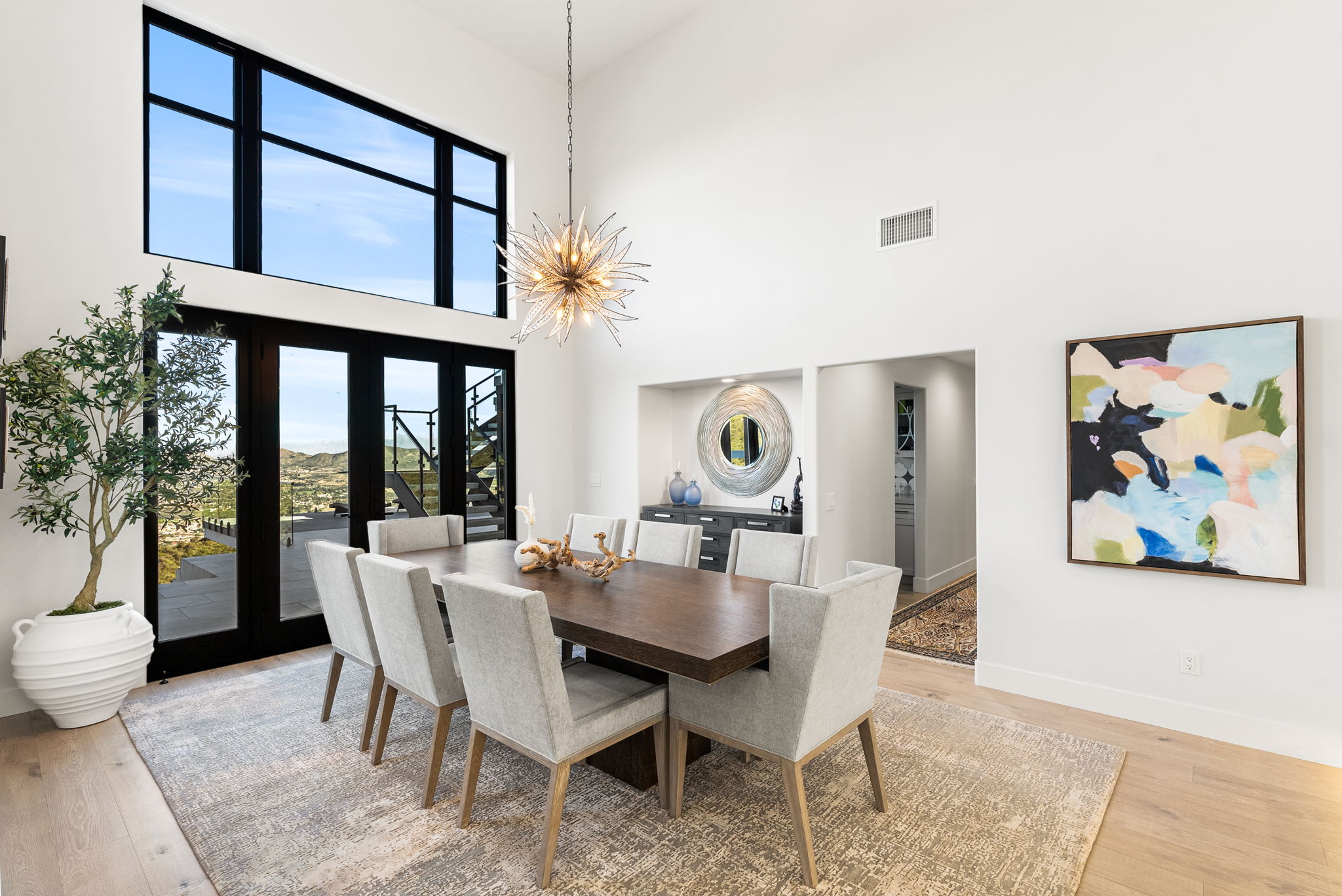 This elegant dining room features a large, dark wood table surrounded by eight upholstered grey chairs, set upon a neutral textured area rug. The space is highlighted by a striking modern starburst chandelier, high ceilings, and floor-to-ceiling windows that provide expansive views and natural light. A piece of abstract art adds a colorful focal point to the clean, white-walled interior, creating a sophisticated and welcoming atmosphere.