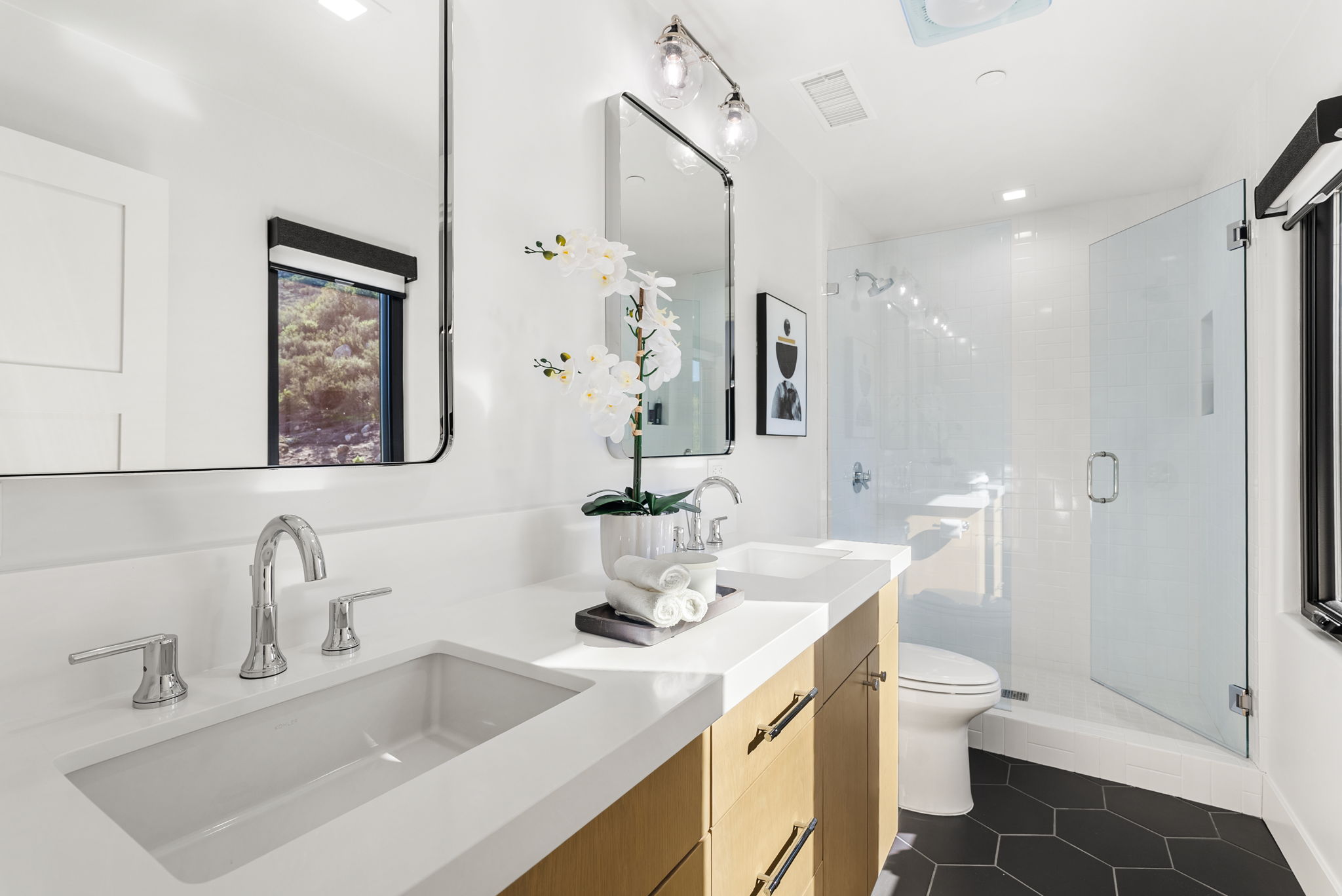 This modern bathroom features a dual-vanity setup with light wood cabinetry, white quartz countertops, and sleek chrome fixtures. The space includes a glass-enclosed walk-in shower, black hexagonal floor tiles, and contemporary mirrors, creating a clean and high-end aesthetic. A window view and a decorative orchid add brightness and a touch of sophistication to the room.