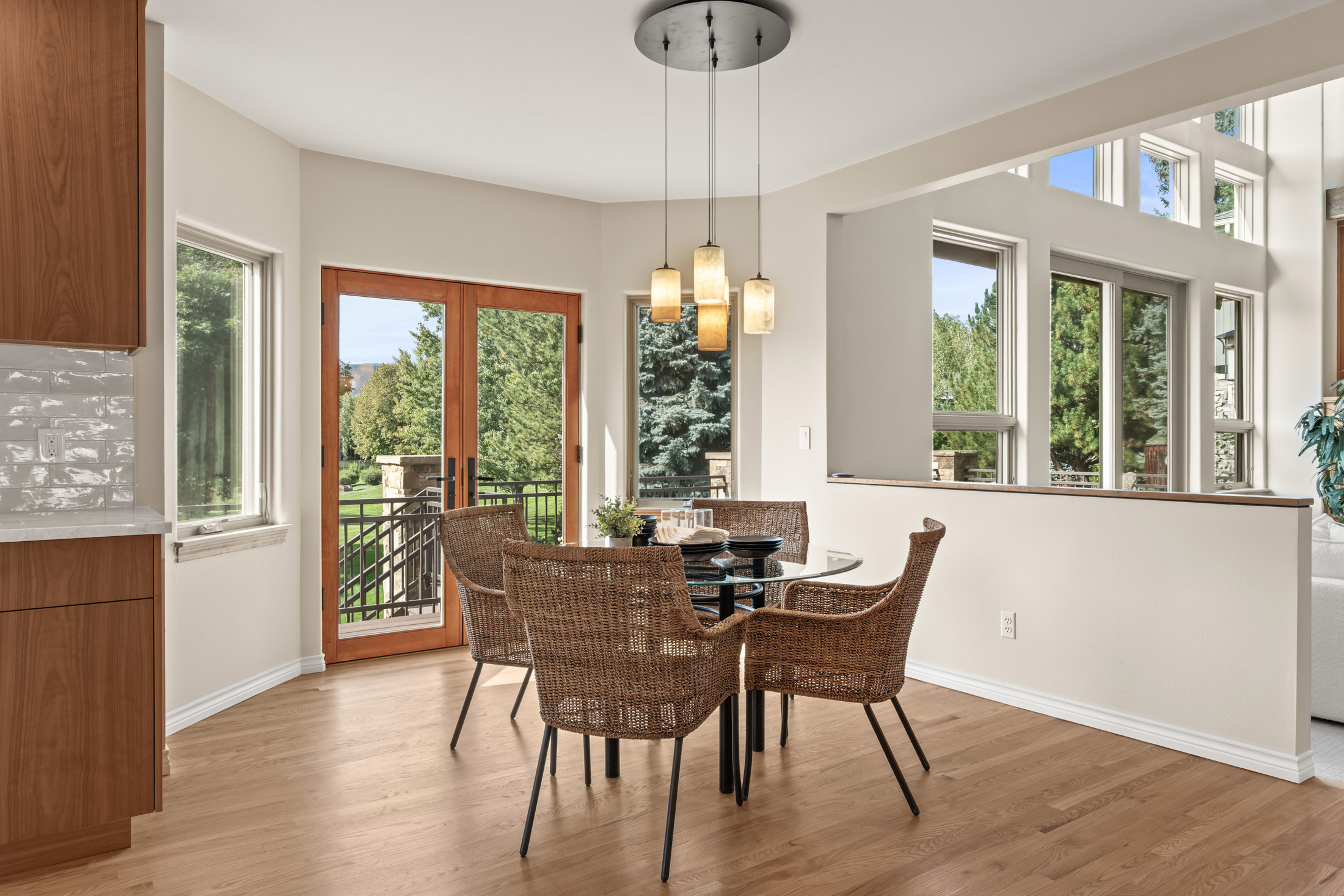 This interior shot showcases a dining area with a round glass table surrounded by four wicker chairs with black metal legs. A modern chandelier hangs above the table, and natural light floods the space through a window and a set of French doors leading to a balcony. The hardwood floors and neutral wall colors create a warm and inviting atmosphere.