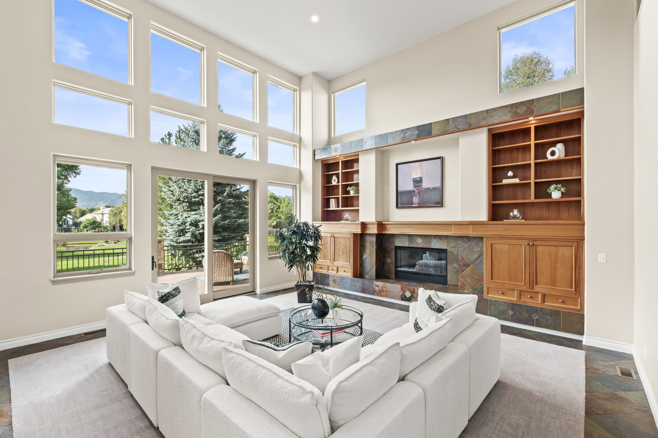 This is a bright and spacious living room featuring a large white sectional sofa, a modern glass coffee table, and a fireplace with built-in shelving. The room is illuminated by natural light streaming through large windows, offering views of the outdoors. The flooring is a combination of carpet and tile, adding texture and visual interest.