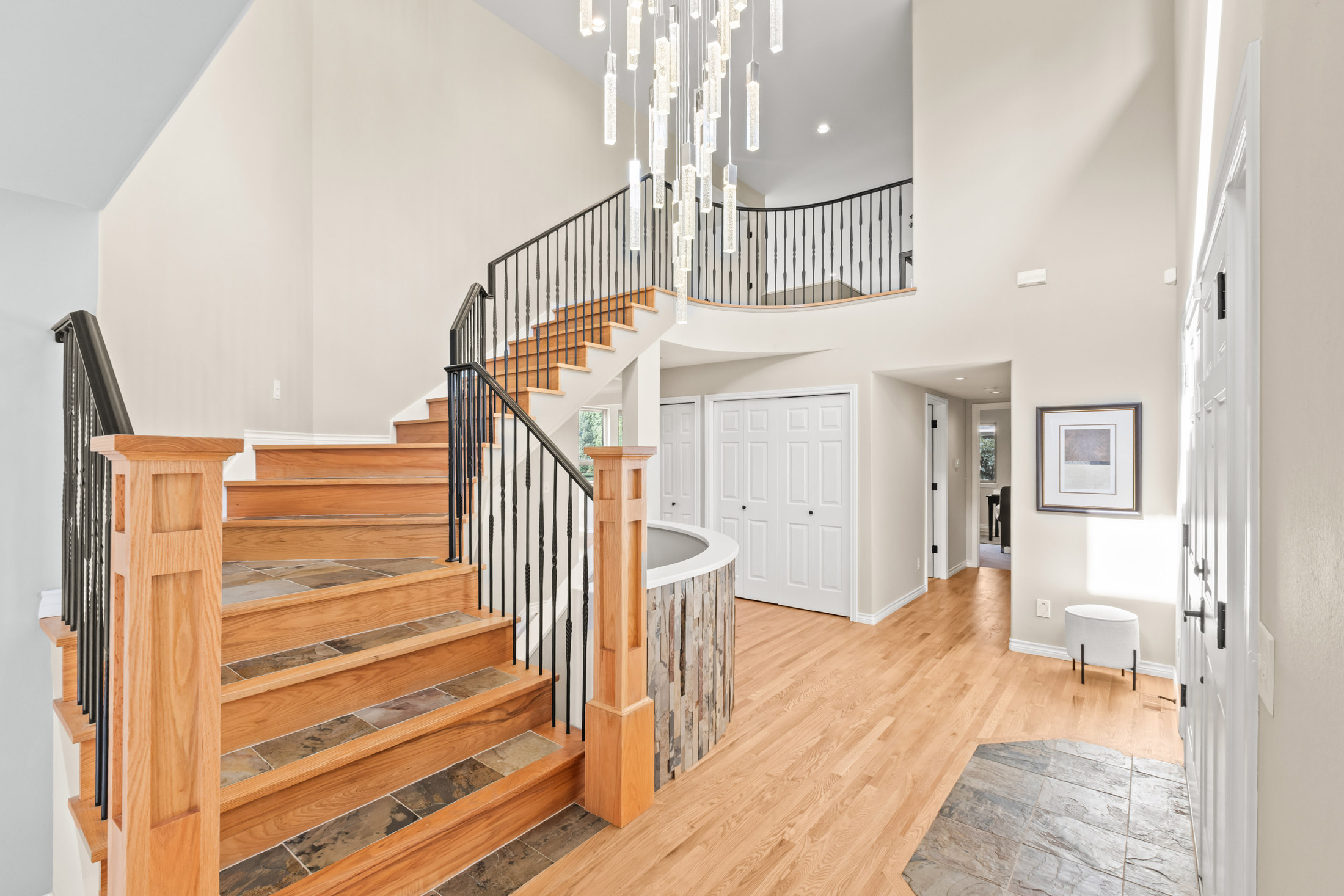 This is an interior shot of a grand foyer featuring a staircase with wooden steps and black wrought iron railings. A modern chandelier hangs from the high ceiling, illuminating the space. The flooring is hardwood, and there are white doors and trim, creating a bright and welcoming entrance.