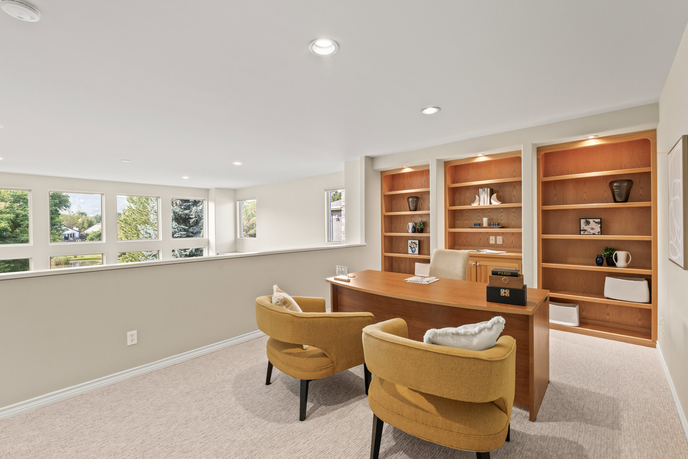 This is an interior shot of a home office featuring a large wooden desk, two mustard-colored armchairs, and built-in wooden bookshelves. The room has a neutral color palette with beige walls and carpet, and natural light streams in through a series of windows. The overall impression is a comfortable and functional workspace.