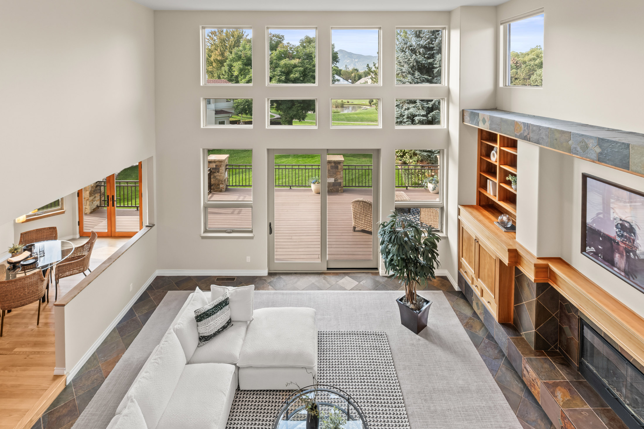 This is a bright and airy living room featuring a soaring two-story wall of windows that offer a view of the outdoor deck and landscape. The room is furnished with a large white sectional sofa, a glass coffee table, and a neutral-toned area rug. A fireplace with a built-in entertainment center adds a cozy focal point to the space.