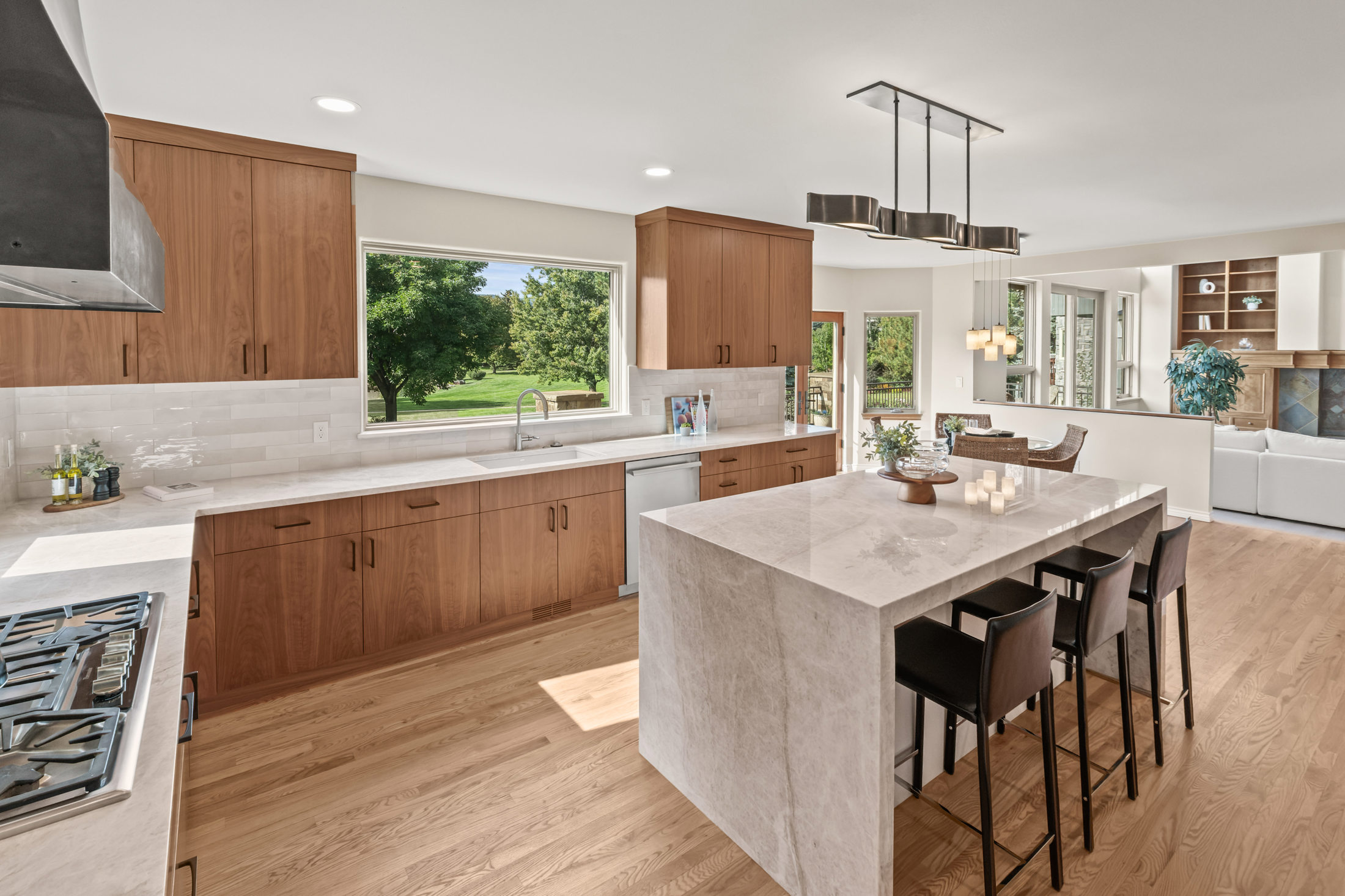 This is a bright and modern kitchen featuring wood cabinetry, white countertops, and a large island with seating. A large window over the sink provides natural light and a view of the outdoors. The kitchen seamlessly flows into the dining and living areas, creating an open and inviting space.