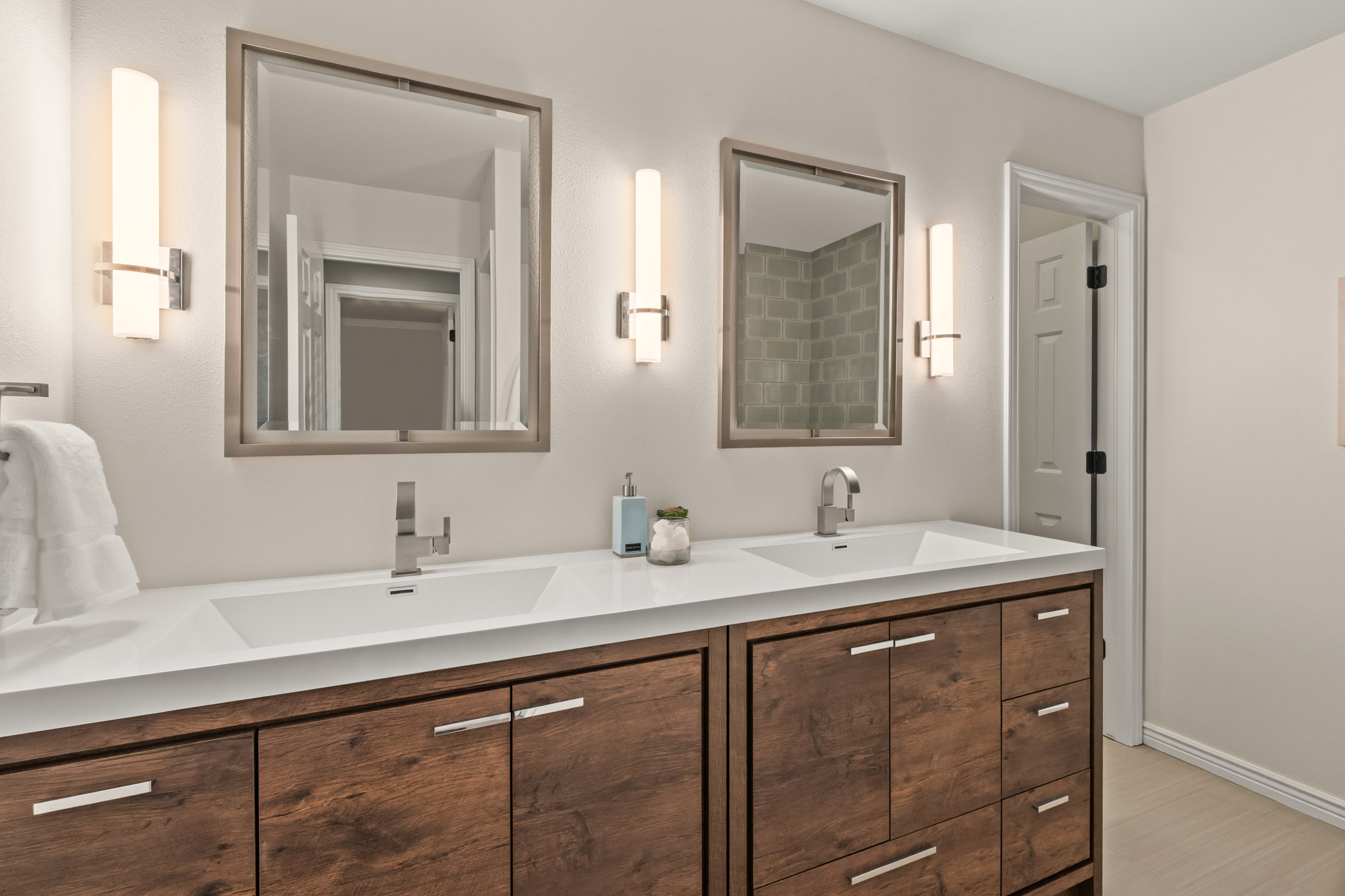 This is a well-lit primary bathroom featuring a double vanity with a white countertop and dark wood cabinets. Each sink has its own mirror and sconce lighting, creating a symmetrical and modern aesthetic. The neutral wall color and clean lines contribute to a spa-like atmosphere.