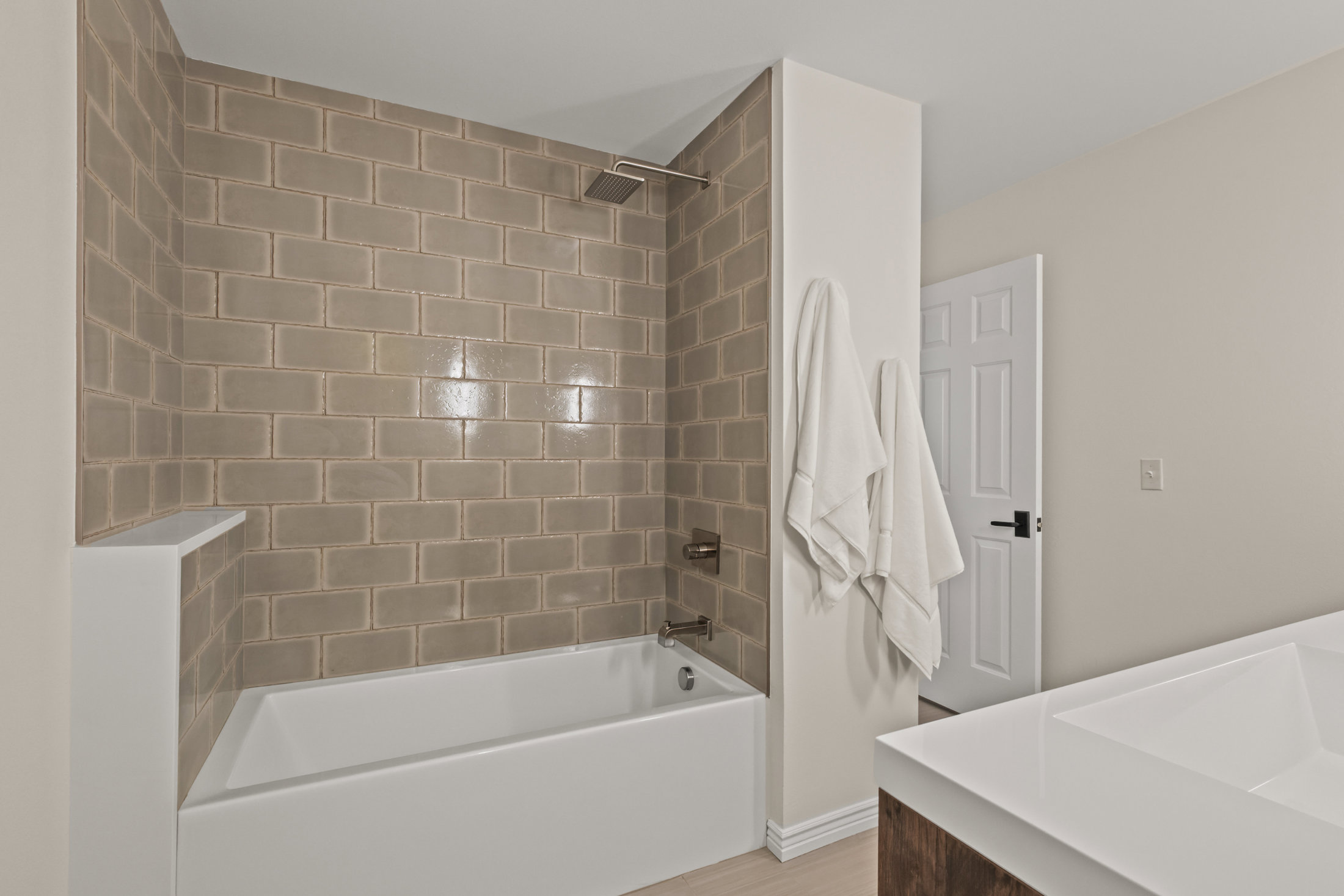 This is a well-lit bathroom featuring a white bathtub with a tiled shower surround in a neutral tone. Two white towels hang on the wall next to the shower. A white door is slightly ajar, and a white countertop with a sink is visible in the foreground, creating a clean and inviting space.