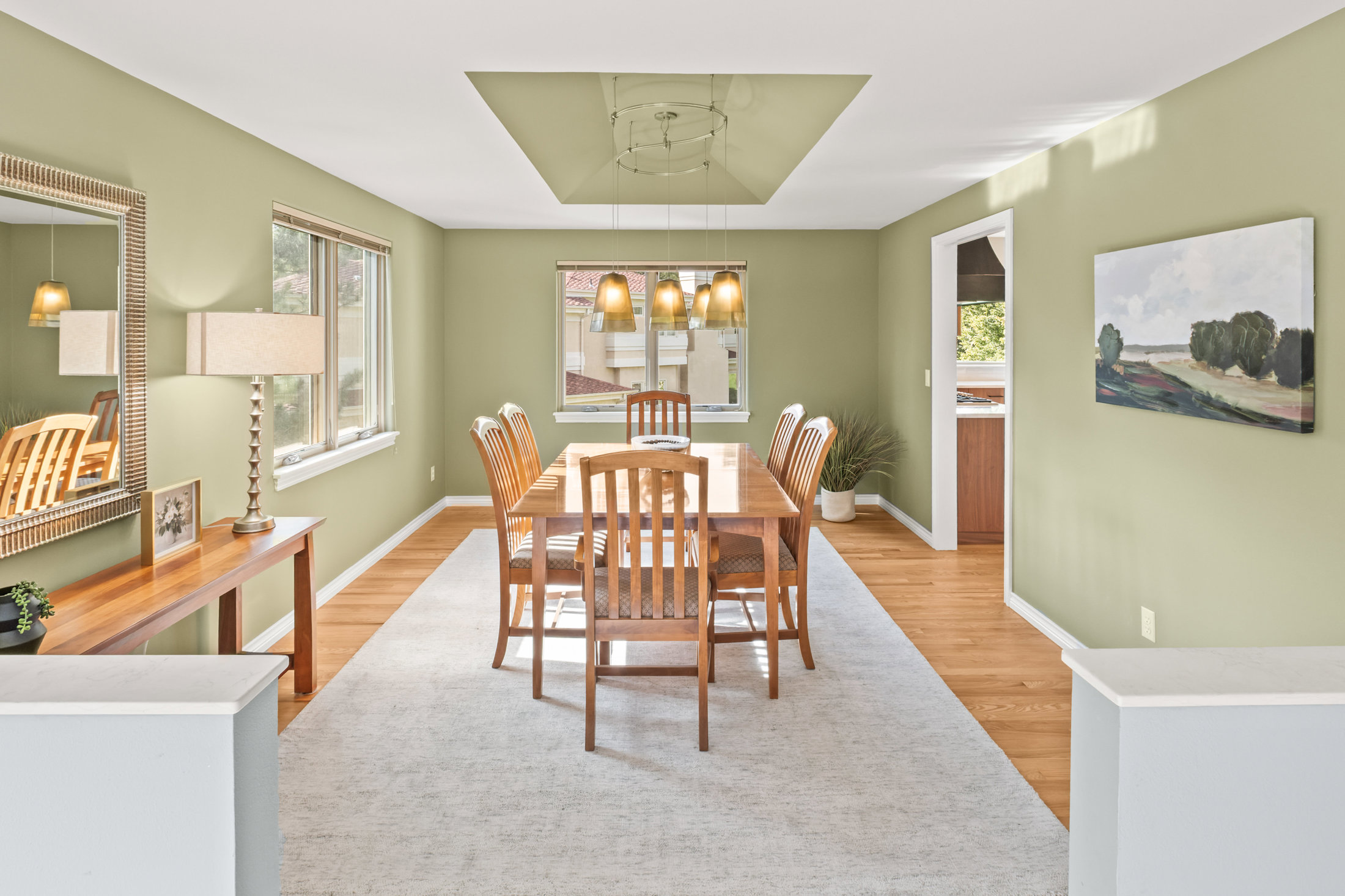This is an interior shot of a dining room featuring a wooden dining table with chairs set on a large area rug. Natural light streams in through the windows, illuminating the light green walls and hardwood floors. A decorative mirror and a landscape painting add to the room's aesthetic appeal, creating a warm and inviting atmosphere.