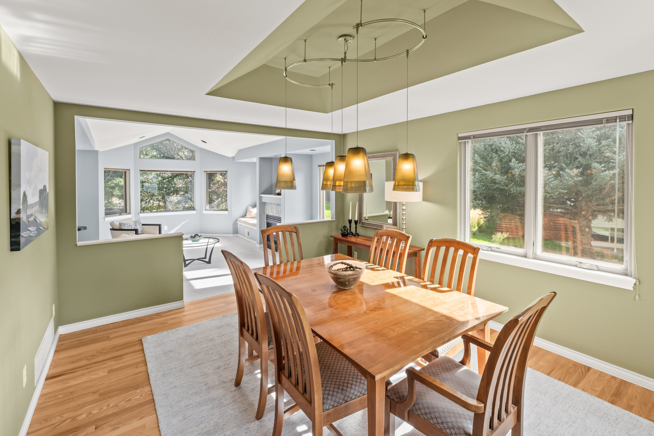 This is an interior shot of a dining room featuring a wooden dining table with six chairs, illuminated by a unique pendant lighting fixture. The room has a warm, inviting feel with light green walls and hardwood floors, complemented by a large window offering natural light and a view of the outdoors. The open layout connects to a living area, enhancing the sense of space and flow.