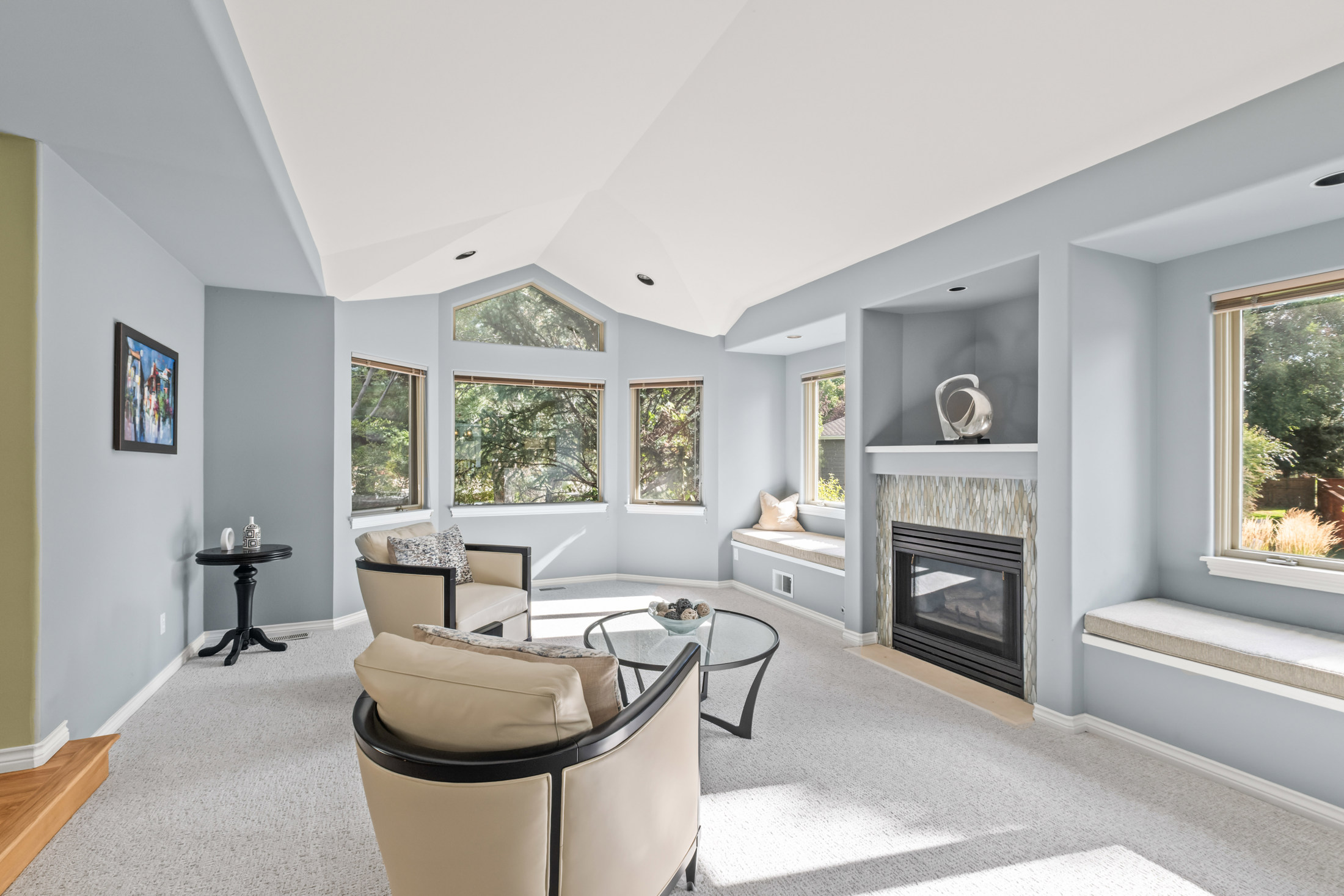 This is a bright and airy living room featuring a vaulted ceiling and multiple windows that provide ample natural light. The room is furnished with two armchairs, a glass coffee table, and a fireplace with a decorative mantel. The light blue walls and neutral carpet create a calming and inviting atmosphere.