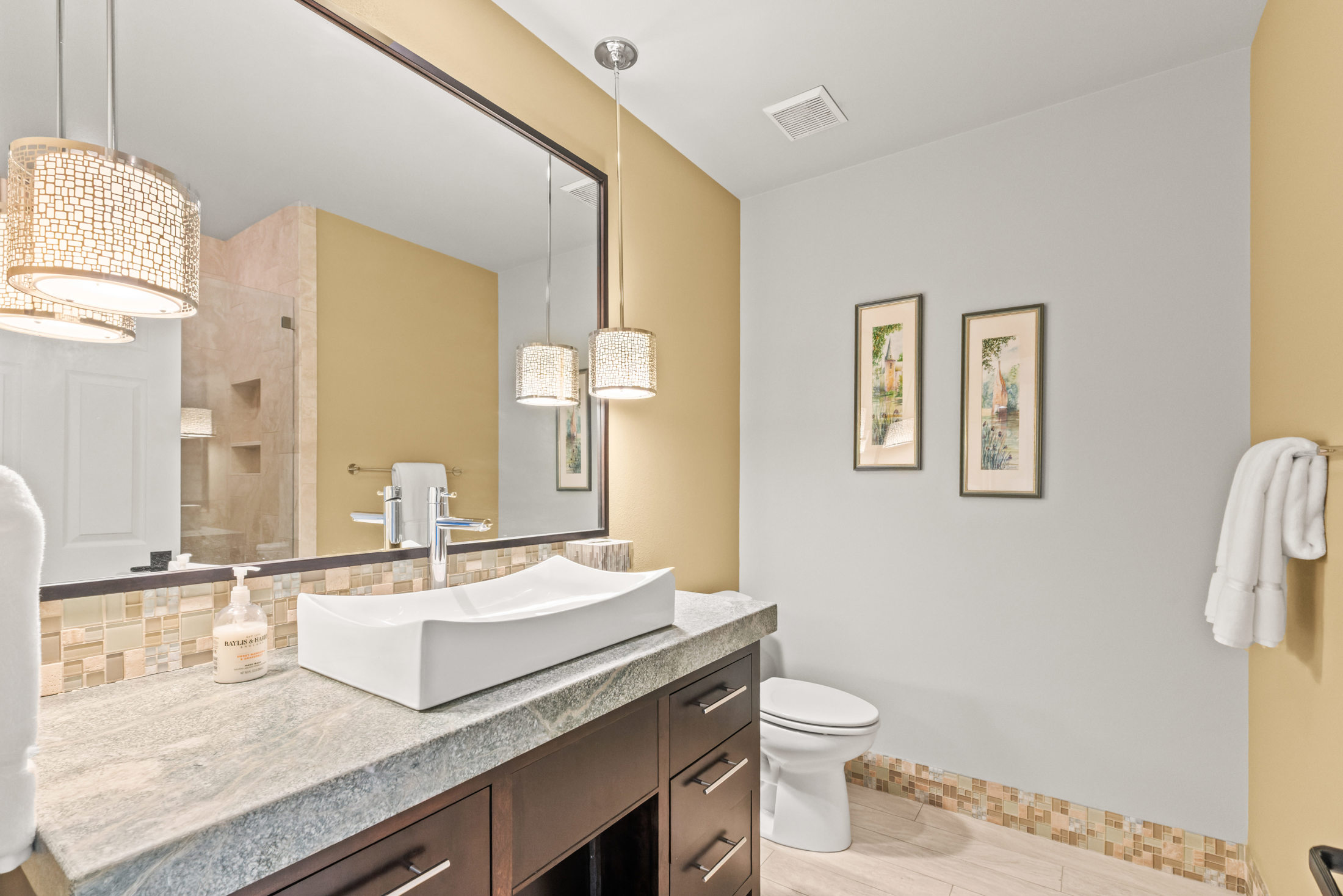This is a well-lit bathroom featuring a modern vessel sink atop a dark wood vanity with a gray countertop. A large mirror reflects the space, including a glimpse of the shower area. The walls are painted in a two-tone scheme, and the flooring consists of light-colored tiles with a decorative mosaic border, creating a clean and inviting atmosphere.