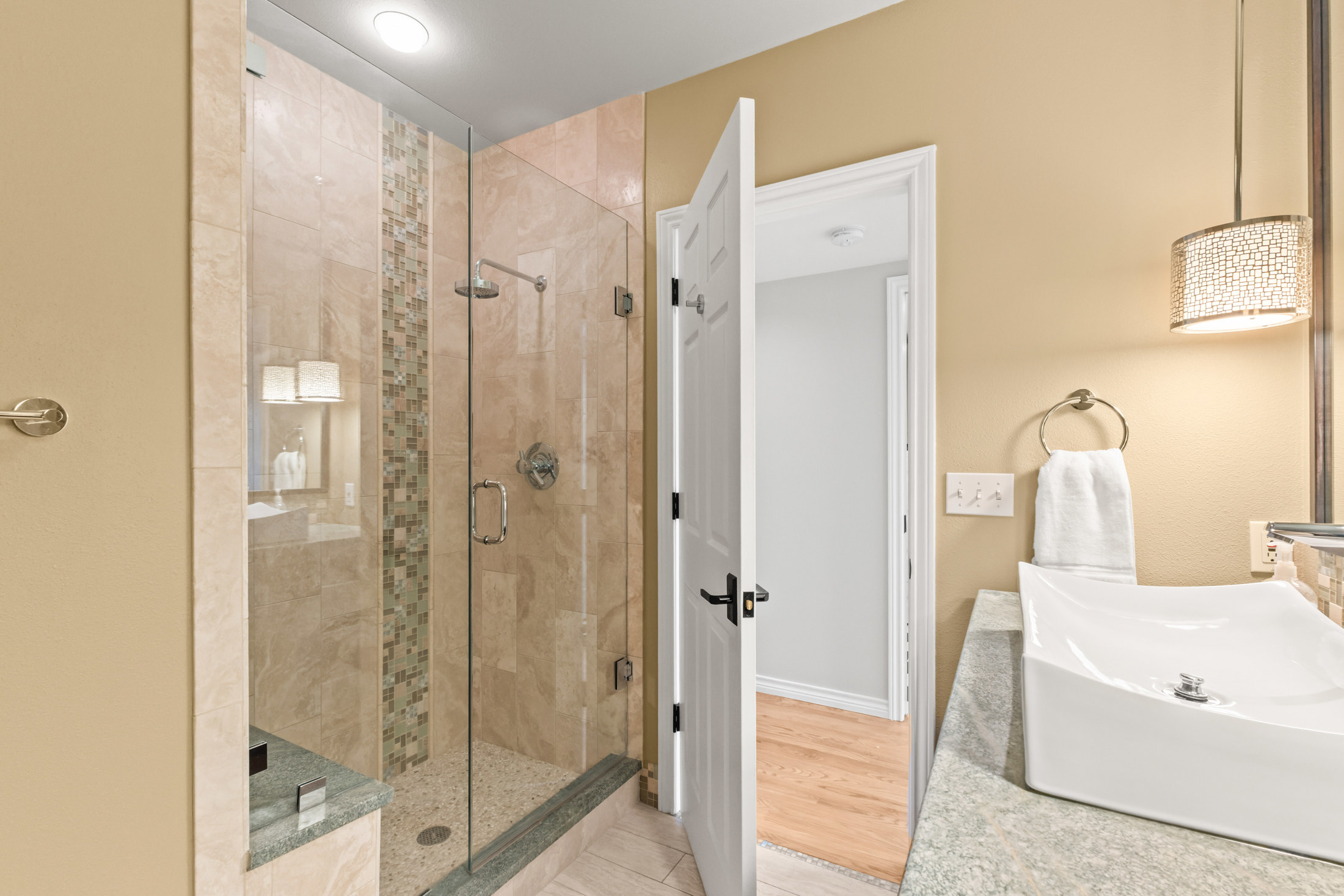 This is a well-lit primary bathroom featuring a glass-enclosed shower with beige tiling and a decorative mosaic strip. The bathroom includes a modern square sink with a granite countertop, a decorative light fixture, and an open doorway leading to another room. The color scheme is warm and inviting, creating a spa-like atmosphere.