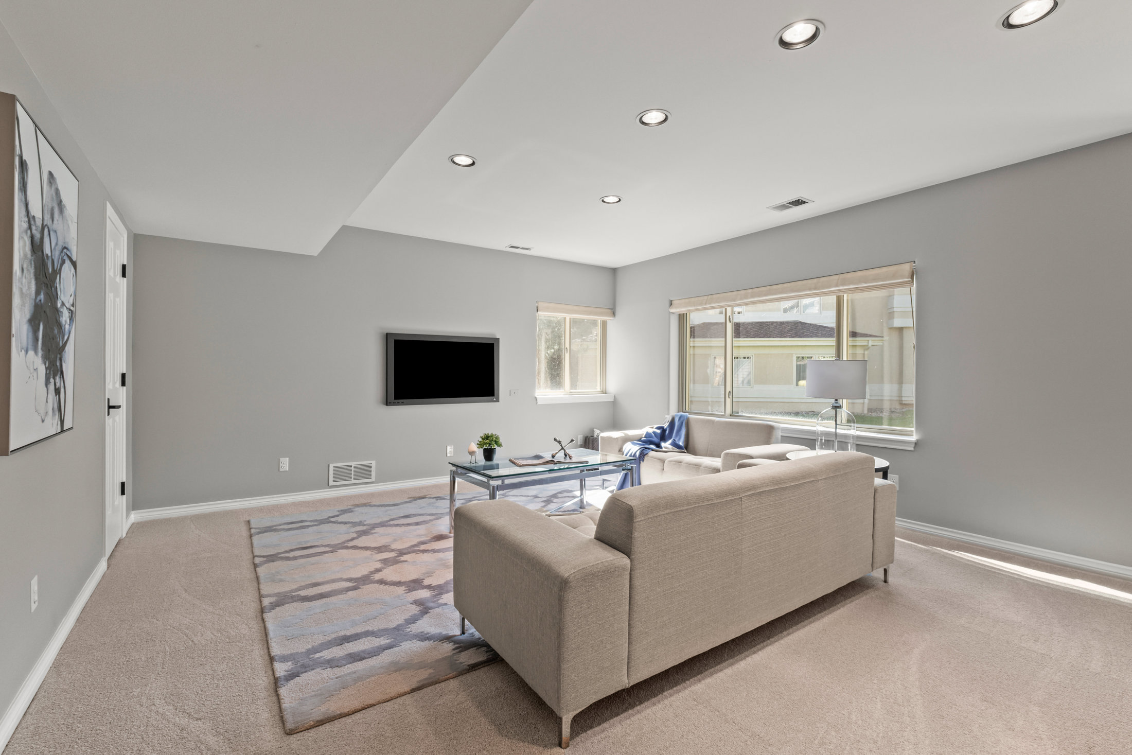 This is an interior shot of a living room featuring a neutral color palette. The room is furnished with a beige sofa, a glass coffee table, and a smaller sofa in the background. A large television is mounted on the wall, and natural light streams in through the windows, creating a bright and inviting atmosphere.