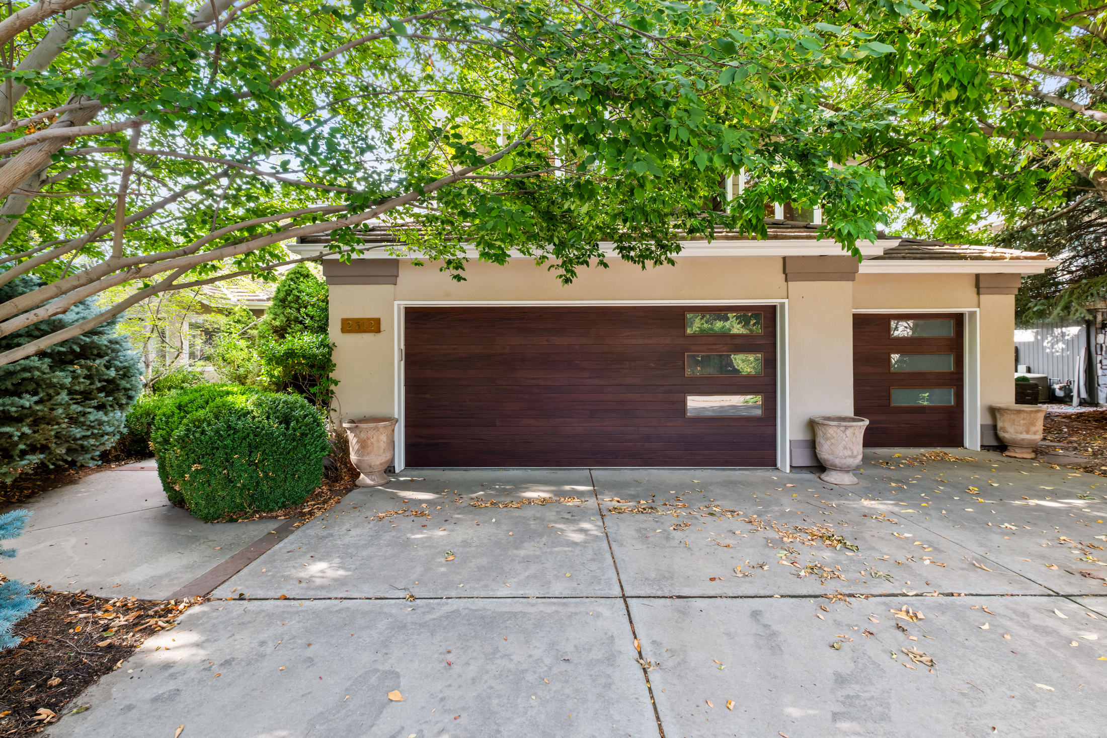 The image showcases a two-car garage with modern wooden doors featuring horizontal panels and rectangular windows. The garage is painted in a neutral beige tone with contrasting trim, and is flanked by decorative urns and lush greenery. The driveway is concrete and shows some fallen leaves, suggesting an established and well-maintained property.