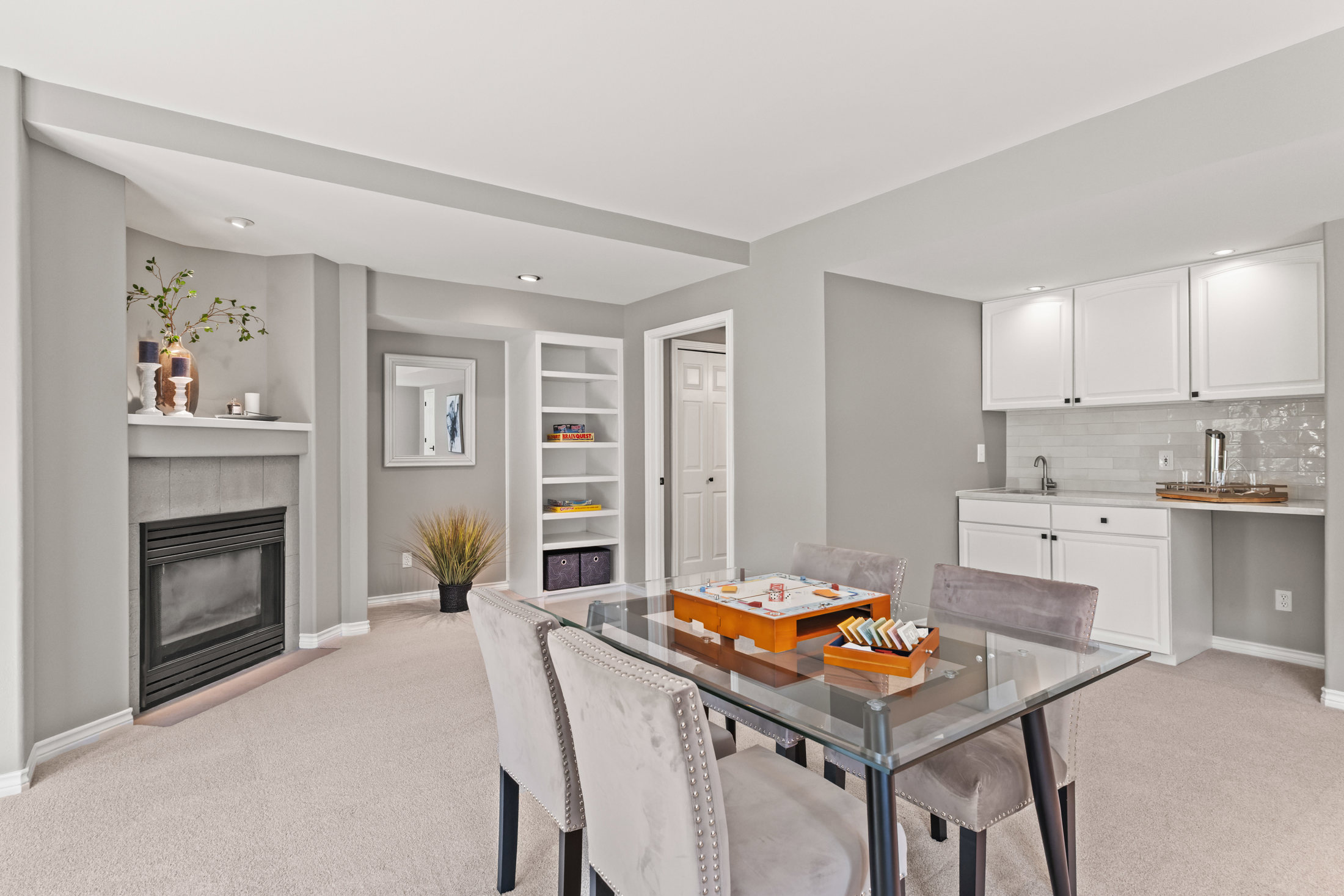 This interior shot showcases a dining room with a modern aesthetic. A glass-topped table with upholstered chairs sits on a neutral carpet, complemented by a built-in bar area with white cabinets and a tiled backsplash. The room also features a fireplace and built-in shelving, contributing to a cozy yet sophisticated atmosphere.