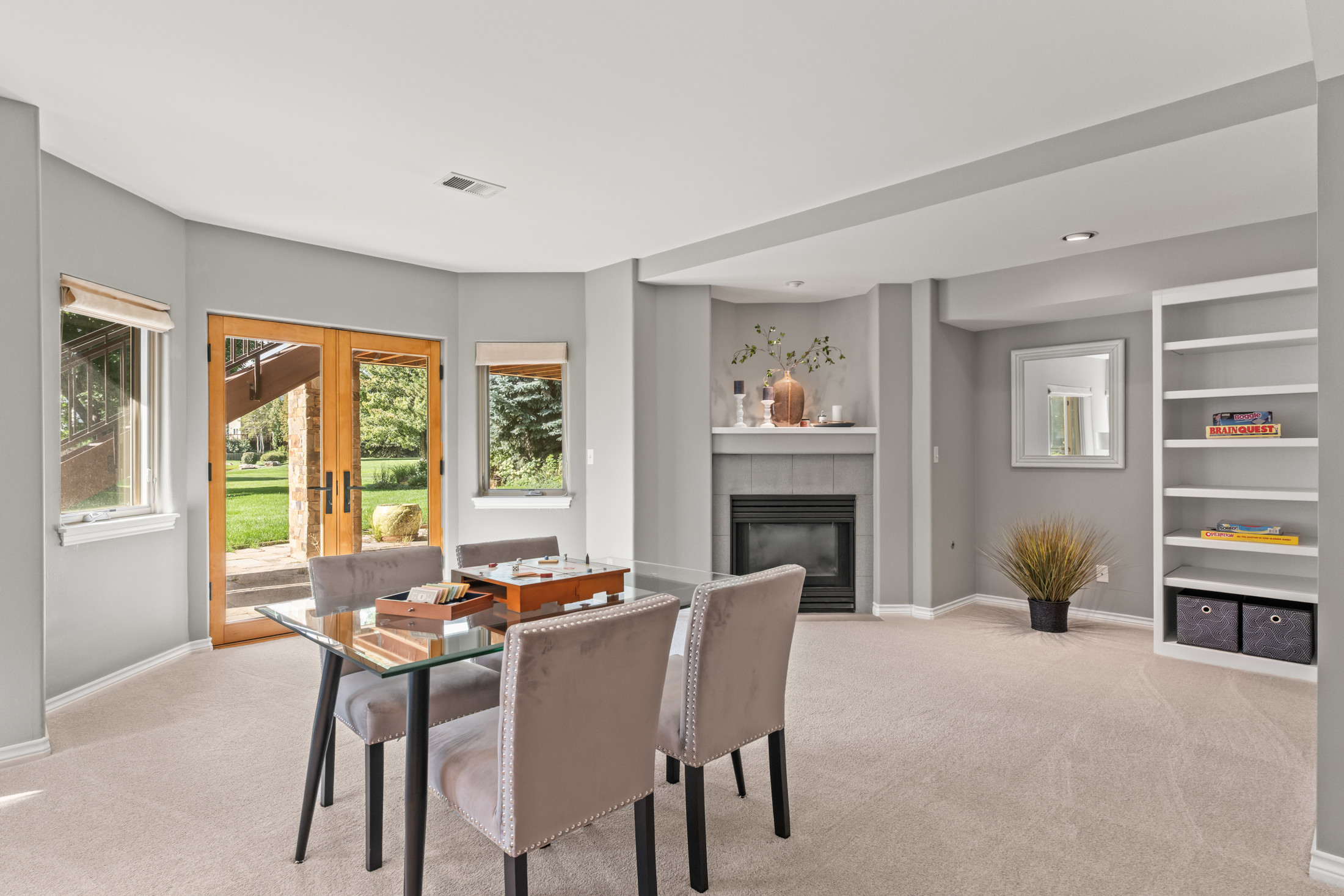 This interior shot showcases a cozy living room with a fireplace and built-in shelving. A glass-topped table with upholstered chairs sits in the foreground, suggesting a game or conversation area. The room is painted in neutral tones with carpet flooring, creating a warm and inviting atmosphere.