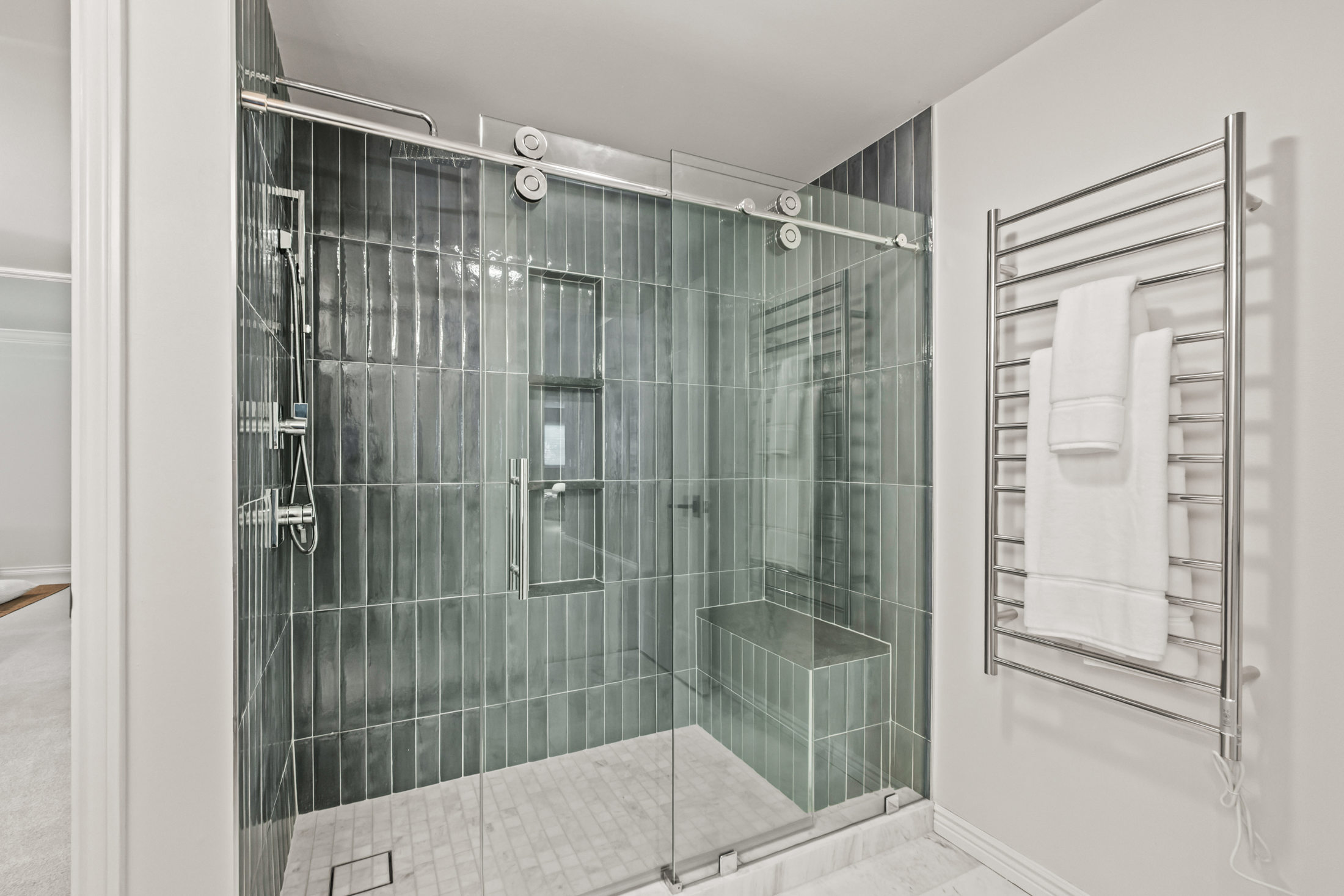 This is a primary bathroom showcasing a modern shower with sleek, vertically stacked gray-green tiles and a frameless glass enclosure. The shower features a built-in niche, a bench, and a contemporary showerhead. A heated towel rack with plush white towels adds a touch of luxury, and the floor is tiled with white marble.