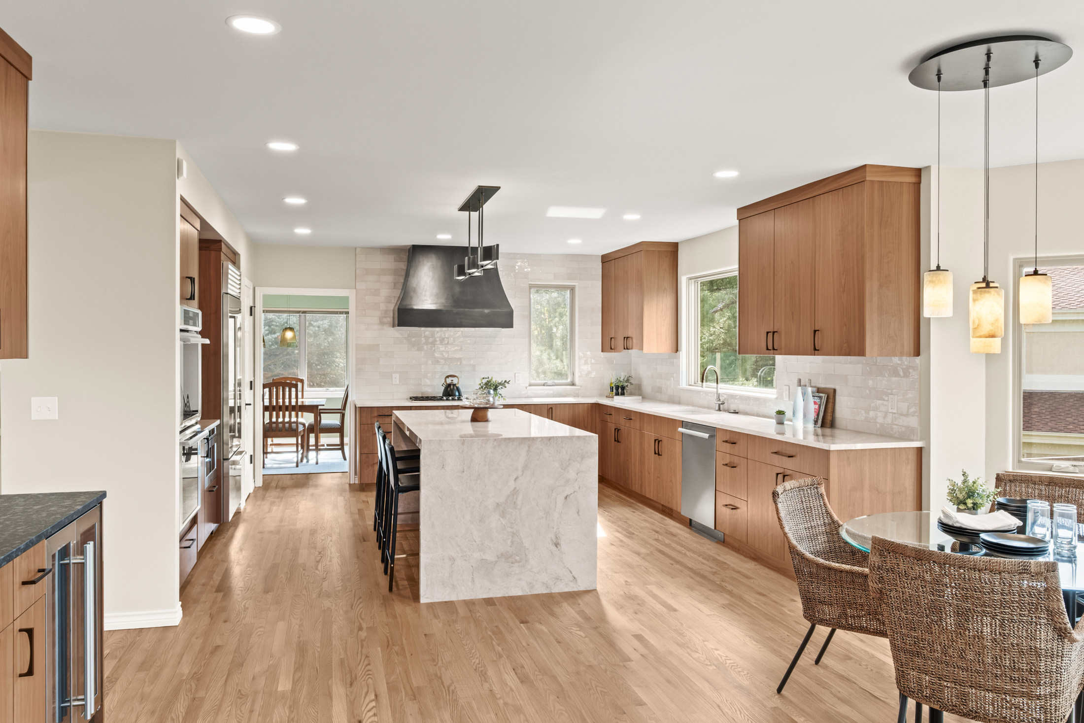 This is a bright and spacious kitchen featuring wood cabinetry, a large marble island with seating, and stainless steel appliances. The kitchen has a modern design with a clean aesthetic, complemented by hardwood floors and ample natural light. A dining area is adjacent to the kitchen, creating an open and inviting space.