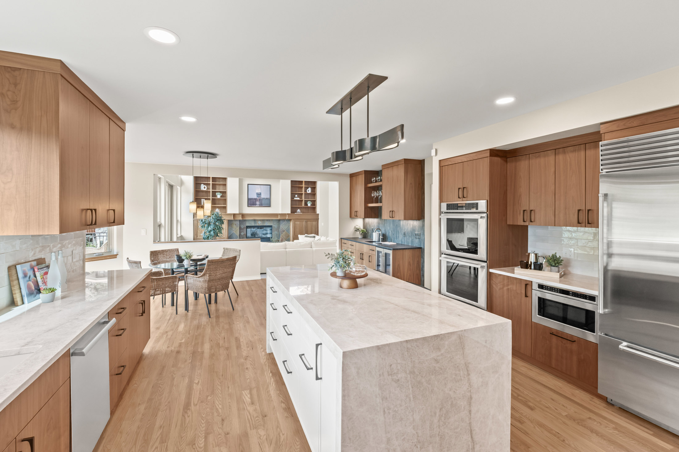 This is a bright and spacious kitchen featuring wood cabinets, stainless steel appliances, and a large island with a marble countertop. The kitchen opens to a dining area and living room, creating an open-concept living space. The hardwood floors and modern lighting fixtures add to the overall appeal of the space.