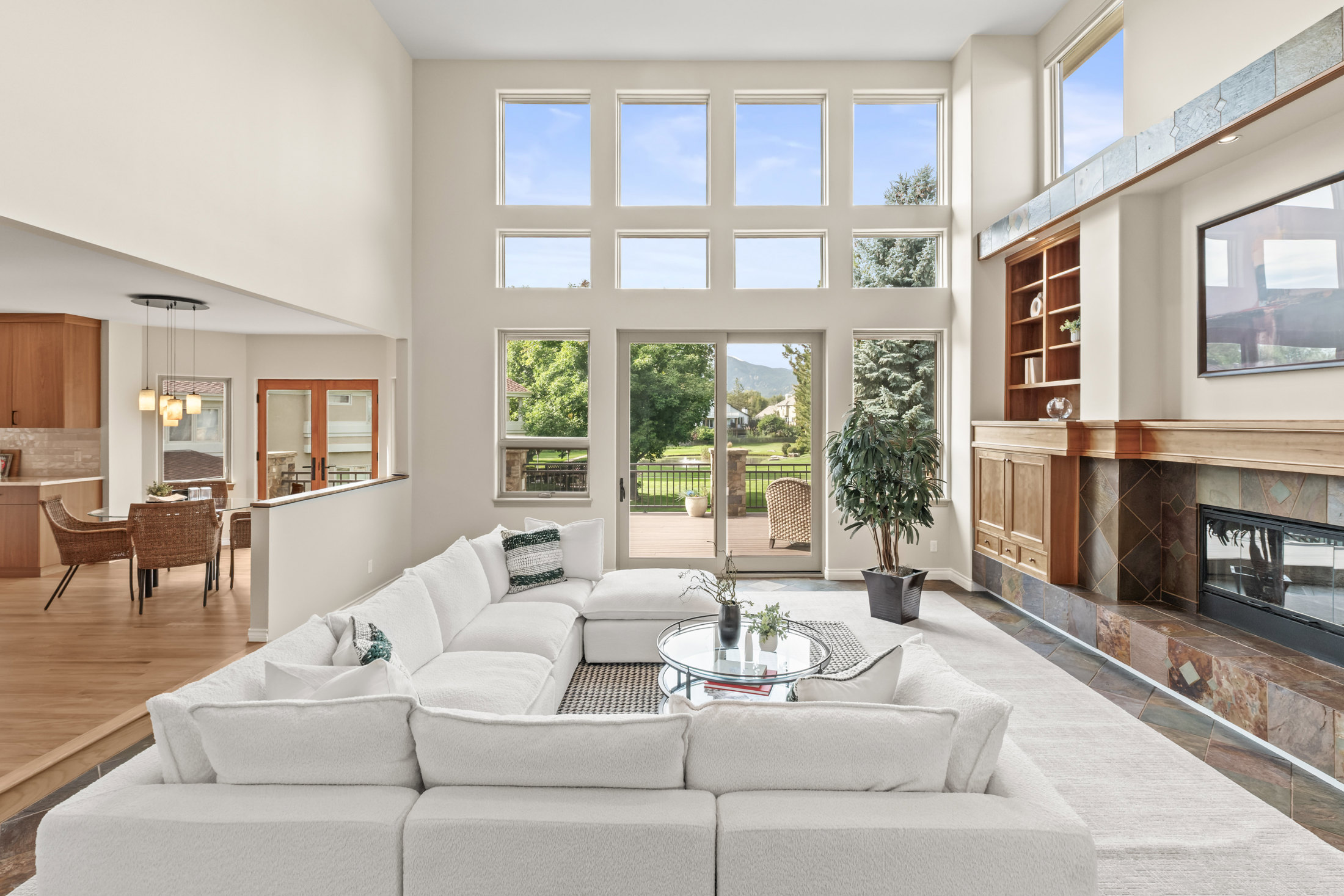 This is a bright and spacious living room featuring a large white sectional sofa, a glass-topped coffee table, and a fireplace with a decorative mantel. The room has high ceilings and large windows that provide ample natural light and views of the outdoor deck and greenery. The open floor plan connects to the dining area and kitchen, creating a seamless flow.