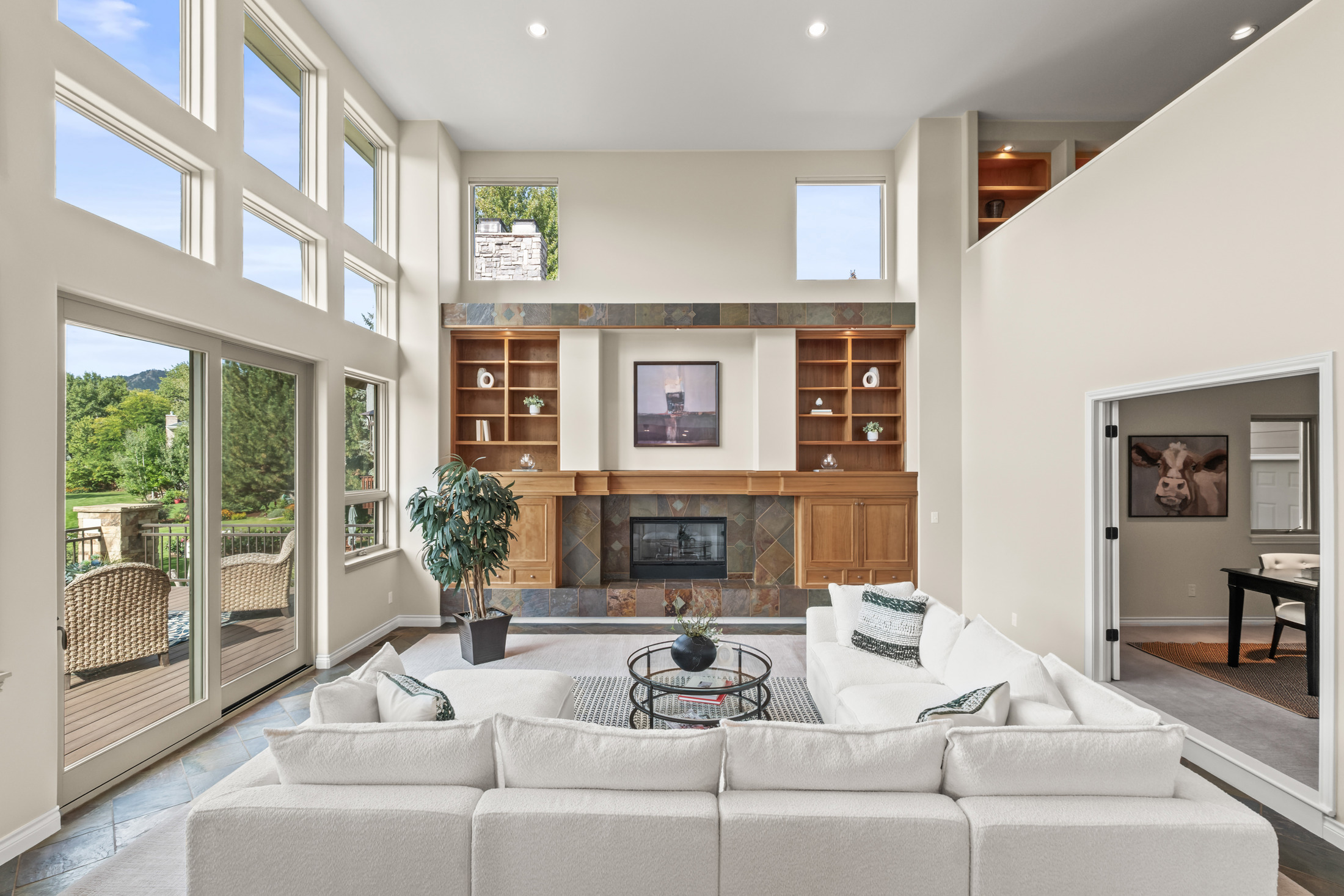 This is a spacious living room featuring a high ceiling with large windows that provide ample natural light and a view of the outdoors. The room is centered around a fireplace with built-in bookshelves on either side. A large, white sectional sofa and a glass-topped coffee table create a comfortable and inviting atmosphere.