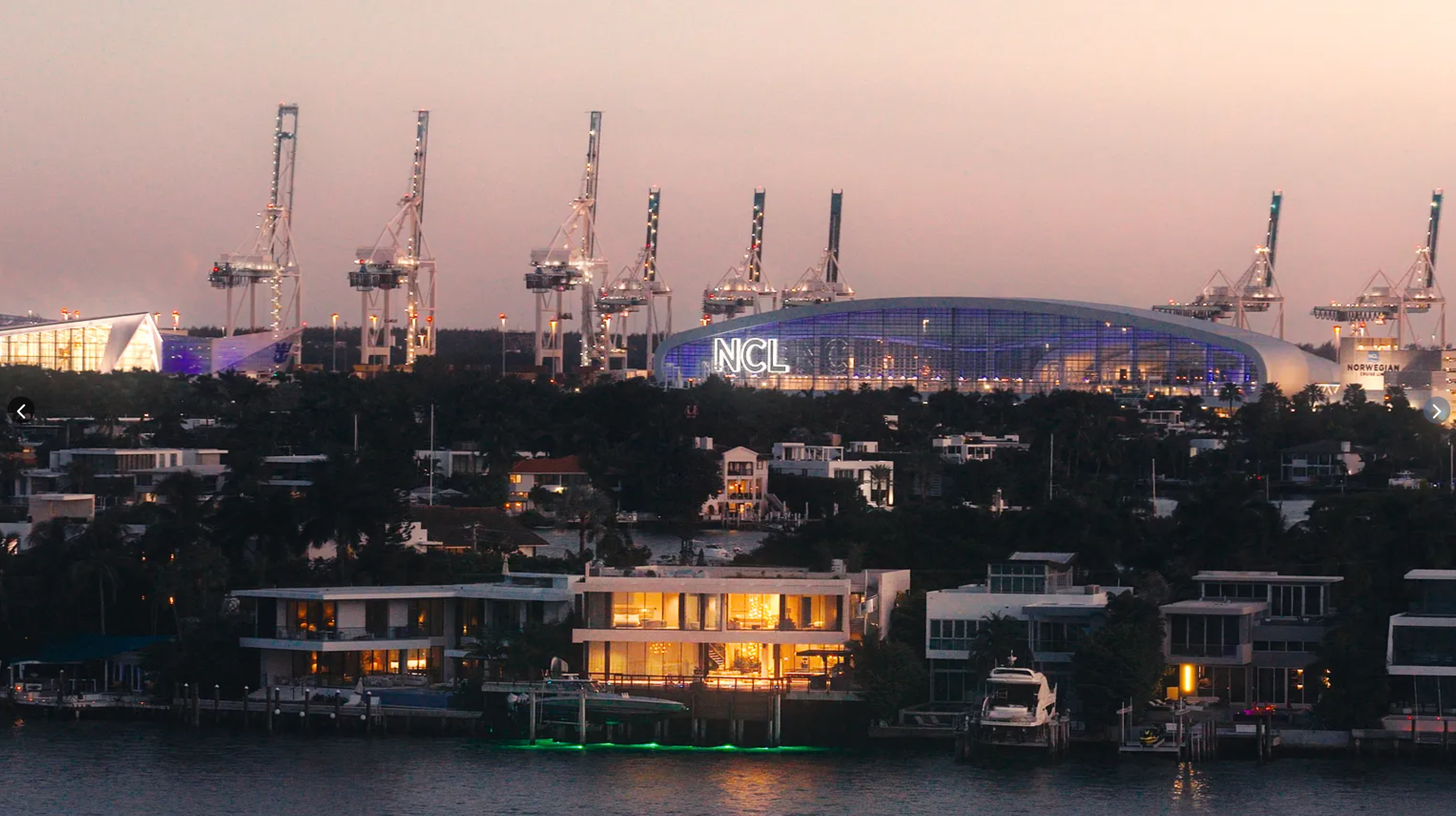 This aerial view showcases a luxury waterfront property at dusk. The modern home features clean lines, large windows, and private docks with boats. In the background, the Miami skyline and port facilities are visible, highlighting the property's prime location and upscale setting.