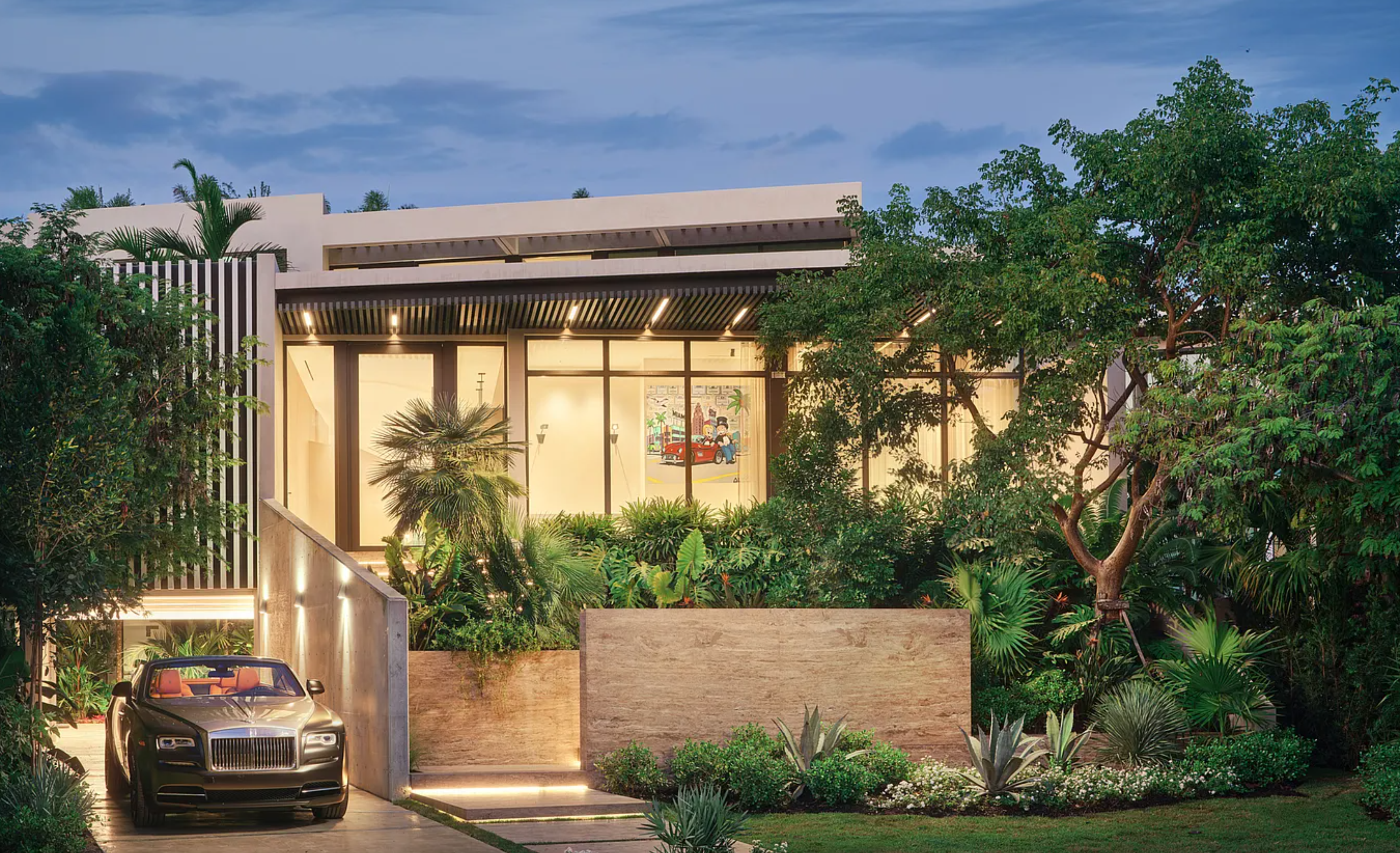 This is a front exterior shot of a modern, luxurious house. The design features large windows, a flat roof with an overhang, and manicured landscaping with trees and greenery. A luxury car is parked in the driveway, adding to the property's high-end appeal.