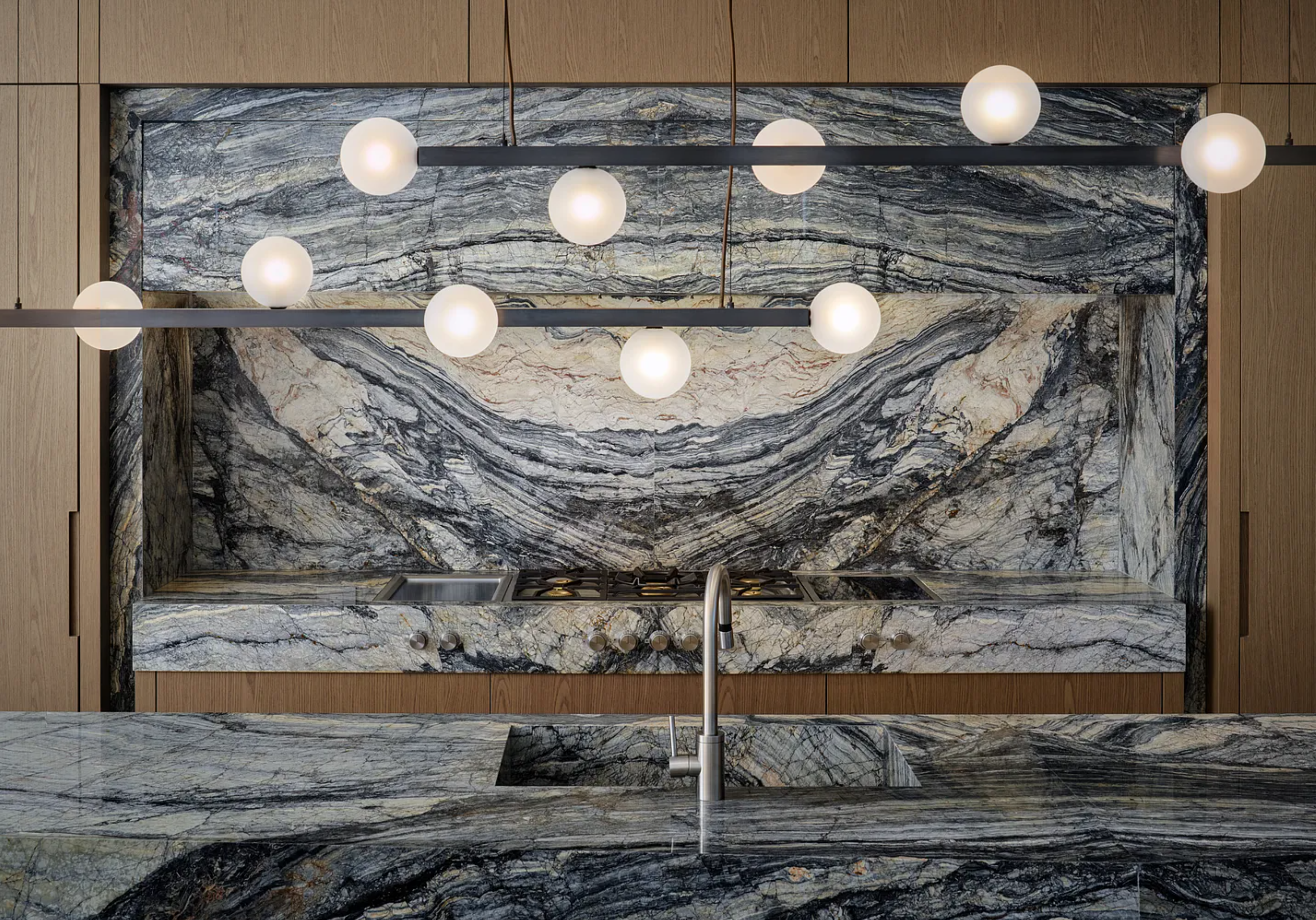 The image showcases a modern kitchen with a striking marble backsplash and countertops featuring a unique, wave-like pattern. A sleek, linear light fixture with globe lights hangs above the island, providing ample illumination. The design emphasizes luxury and sophistication, making it a focal point of the home.