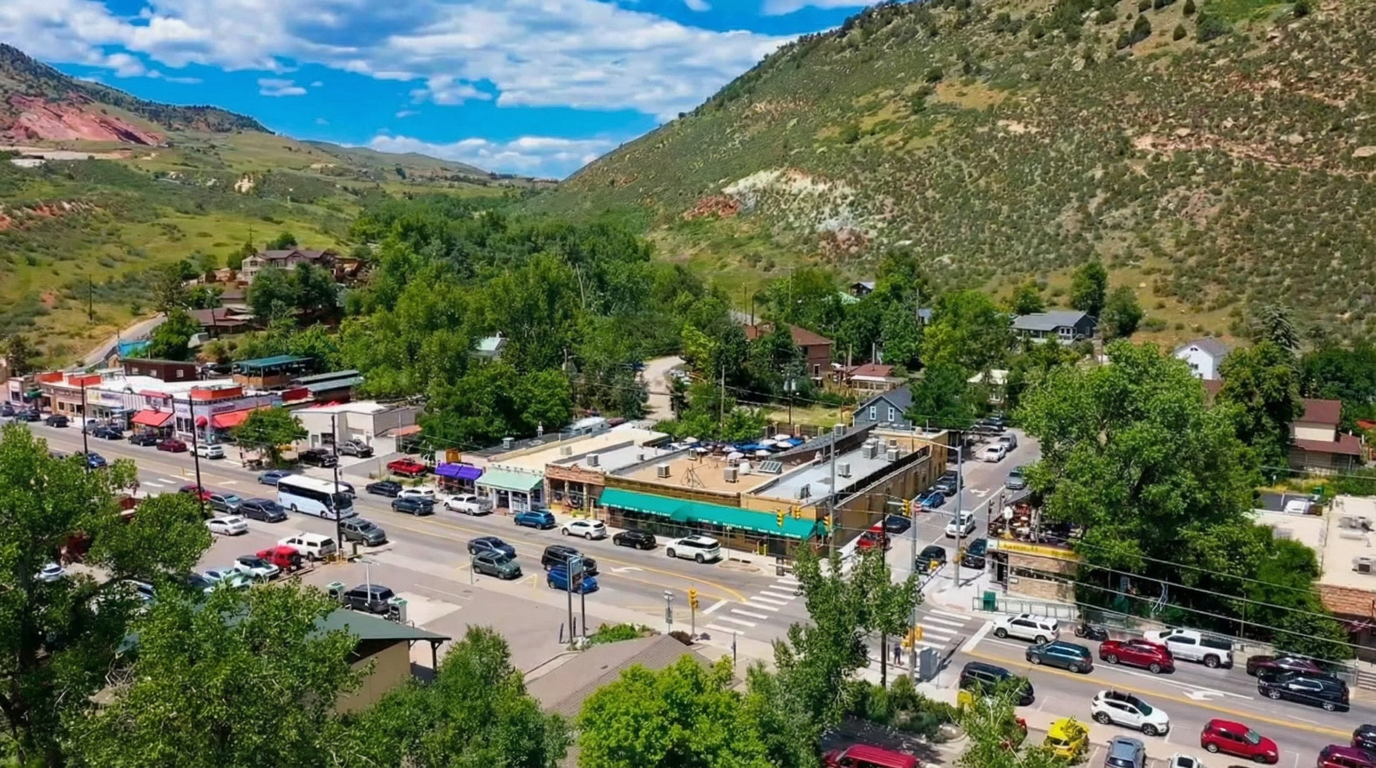 This aerial shot captures a bustling town nestled in a valley, surrounded by green mountains. Buildings are visible with colorful awnings and rooftop features, and a main street is filled with vehicles and pedestrians. The image showcases a vibrant community with a picturesque natural backdrop, making it an attractive location.