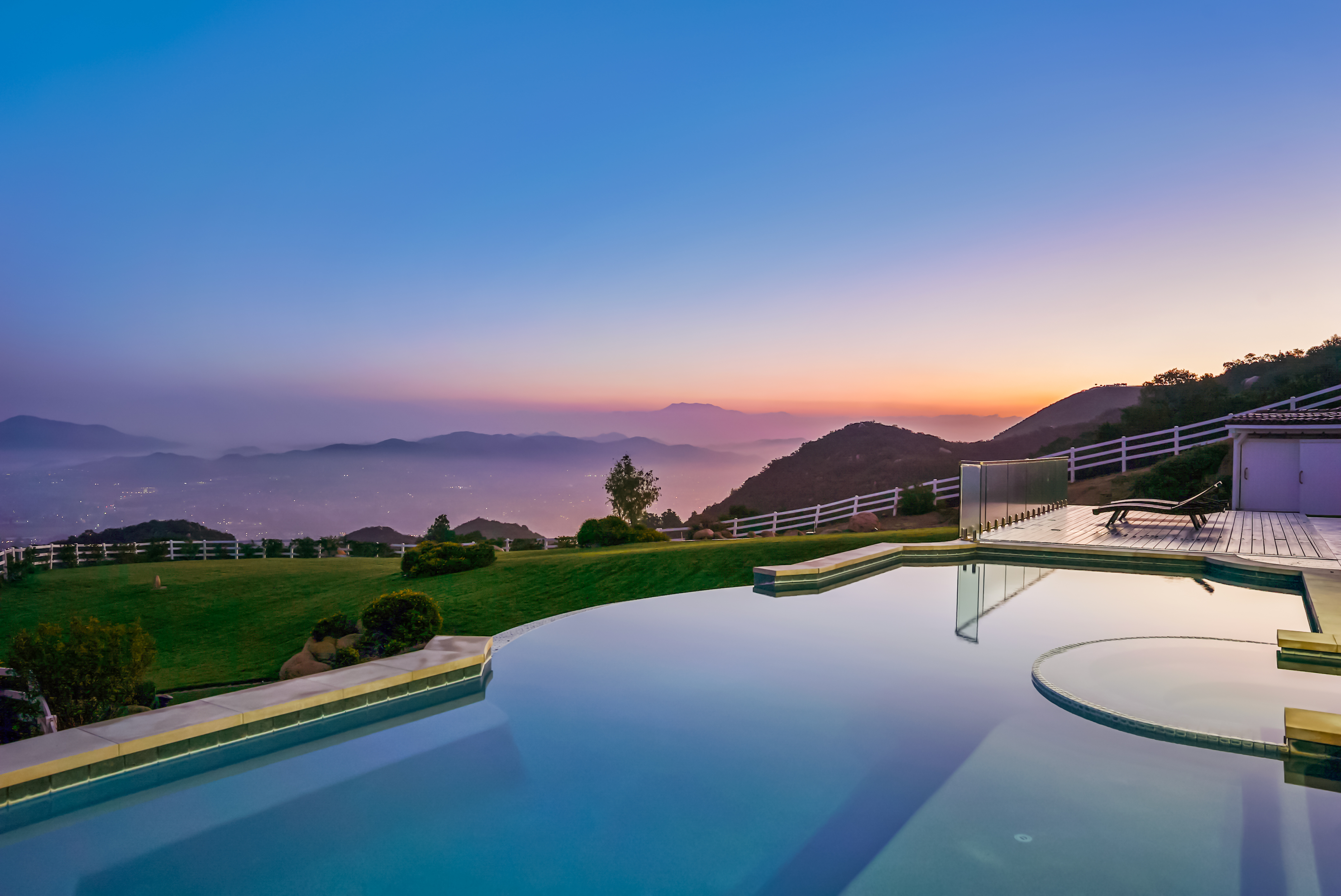 This stunning infinity-edge pool overlooks a vast, mountainous landscape during a serene sunset. The scene features a well-manicured lawn, a wooden deck with lounge chairs, and a glass-railed perimeter that enhances the panoramic view. The composition captures a luxurious, tranquil atmosphere, emphasizing the seamless integration between the property's outdoor amenities and the natural horizon.