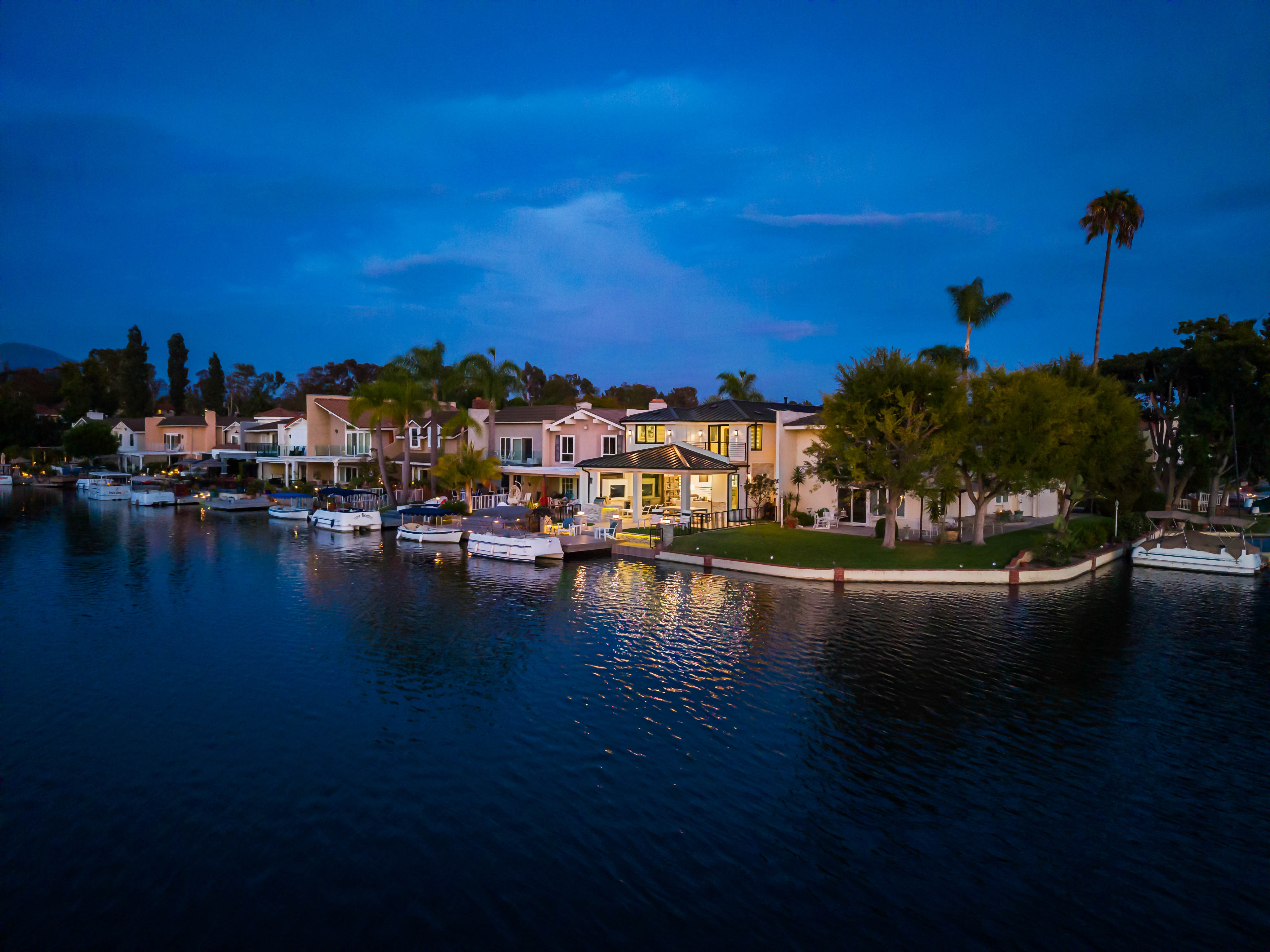 This stunning aerial view captures a luxurious waterfront estate at twilight, showcasing its prime location on a calm canal. The property features a spacious, illuminated outdoor living area with a covered patio that seamlessly transitions to a private dock lined with multiple boats. Lush landscaping, mature trees, and a manicured lawn frame the residence, creating an inviting and serene atmosphere perfect for evening relaxation.