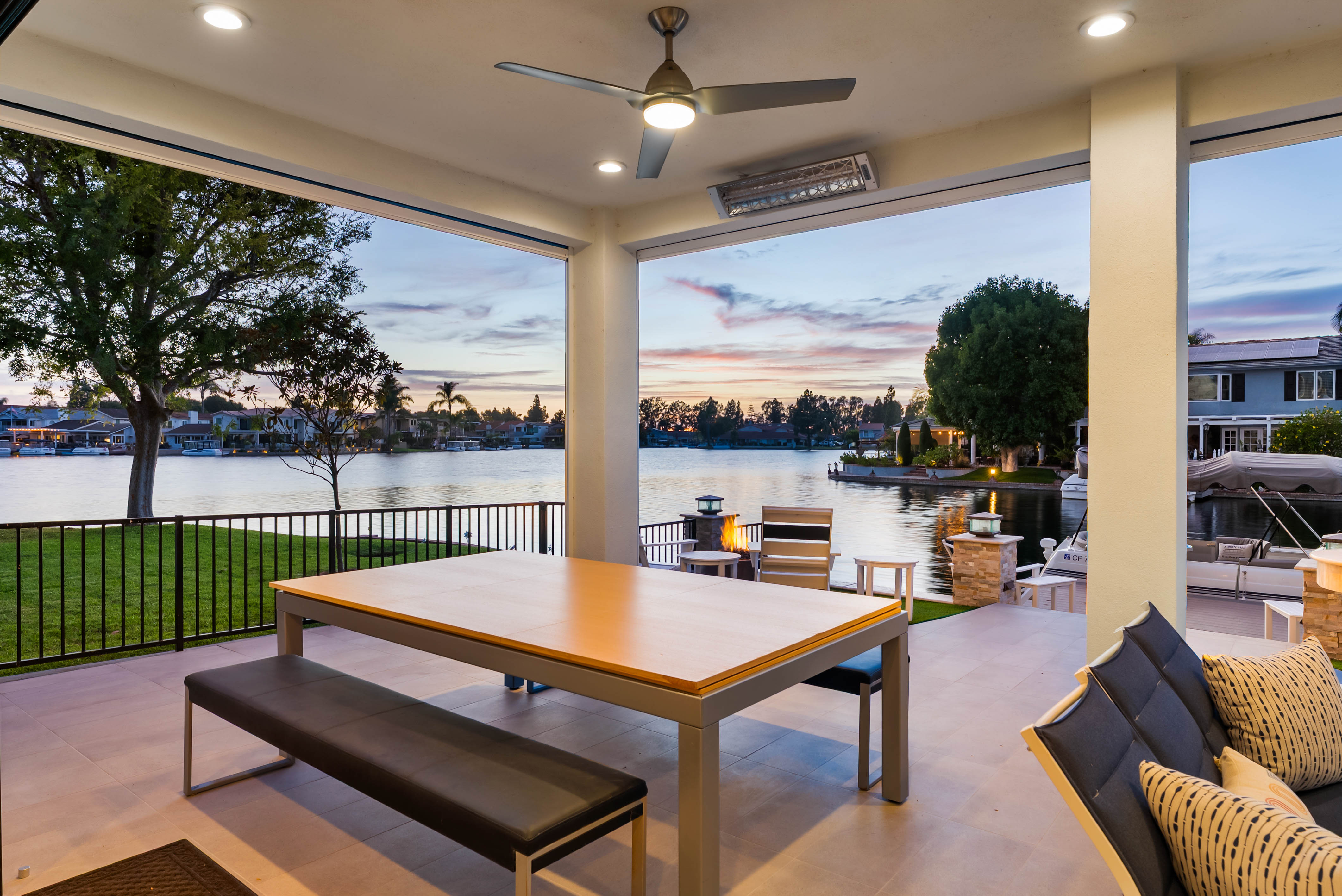 This covered outdoor patio offers a serene, open-air living space overlooking a tranquil lake at sunset. The area is furnished with a modern dining table and bench seating, complemented by comfortable lounge chairs and a small fire feature near the water's edge. The perspective captures a seamless transition between the indoor-outdoor living space and the picturesque waterfront scenery.