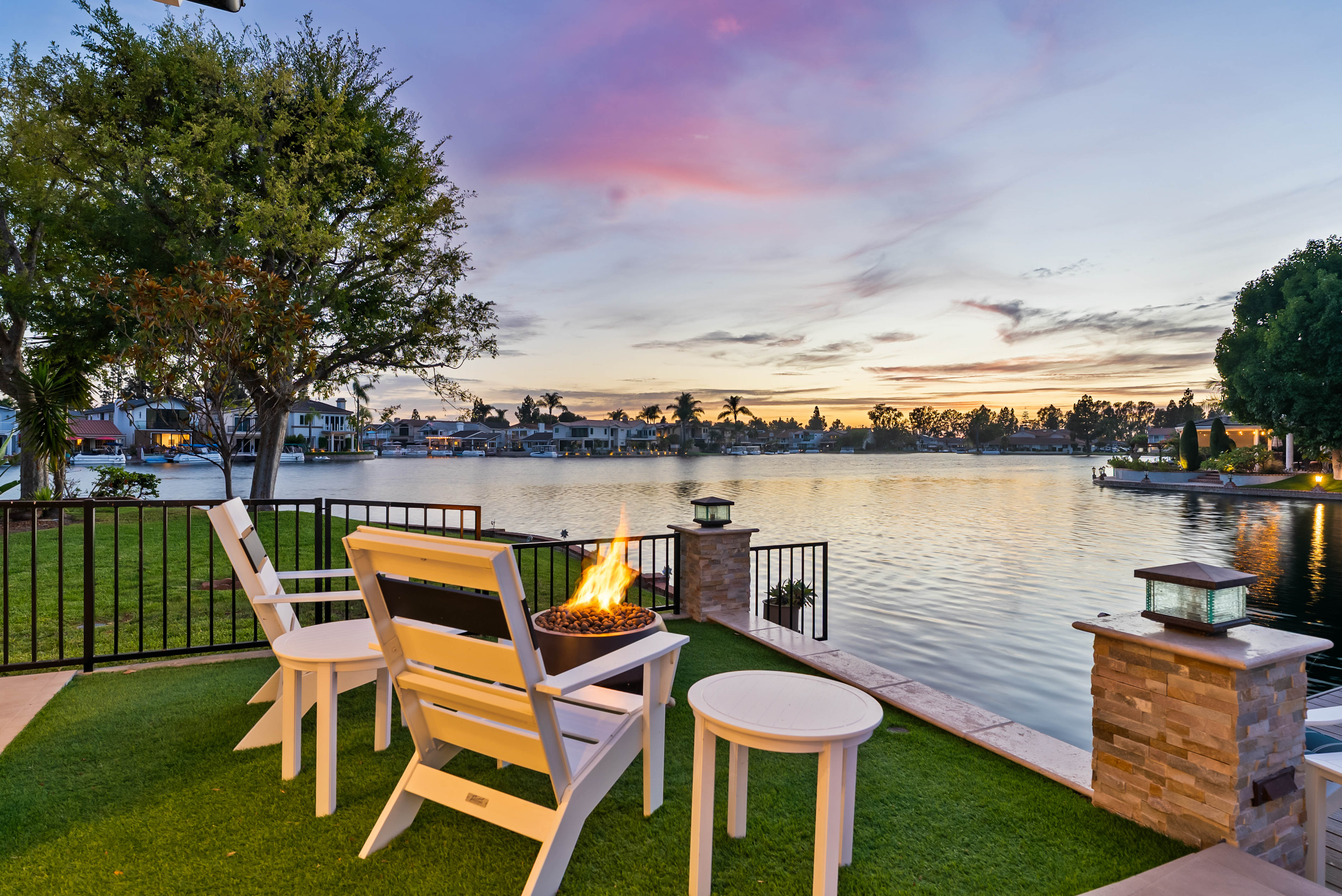 This serene waterfront patio features a pair of white Adirondack chairs positioned around a circular fire pit, creating an inviting space for relaxation. The area is finished with artificial turf and bordered by a black metal fence, overlooking a calm lake at sunset with a vibrant, colorful sky. The scene is framed by stone-capped pillars with integrated lighting, emphasizing a peaceful and luxurious outdoor living environment.