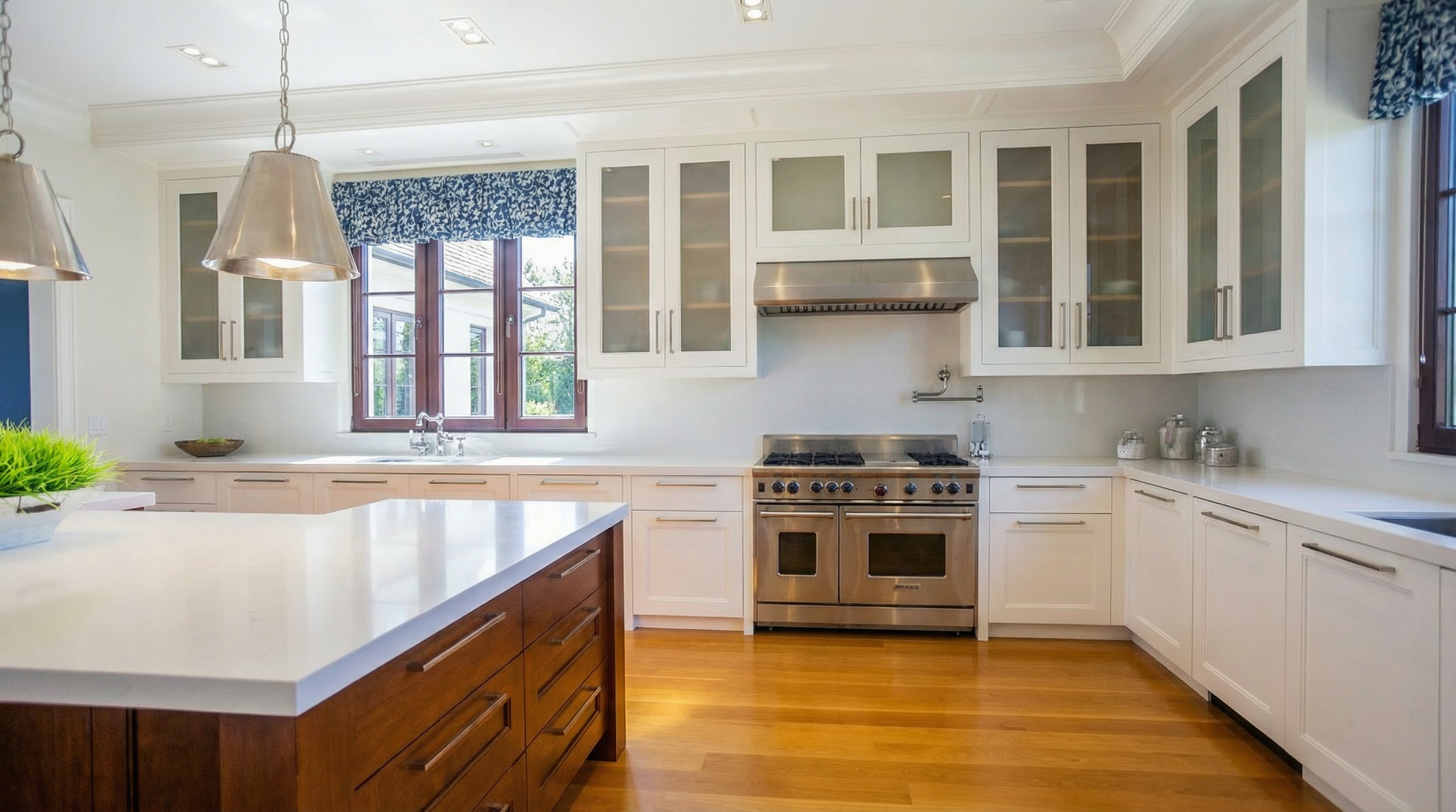 This is a well-lit kitchen interior featuring white cabinetry, a stainless steel range with double ovens, and a wooden kitchen island topped with a white countertop. The hardwood floors add warmth to the space, while the glass-front cabinets contribute to a bright and airy ambiance. The window above the sink offers a view to the outside adding natural light.