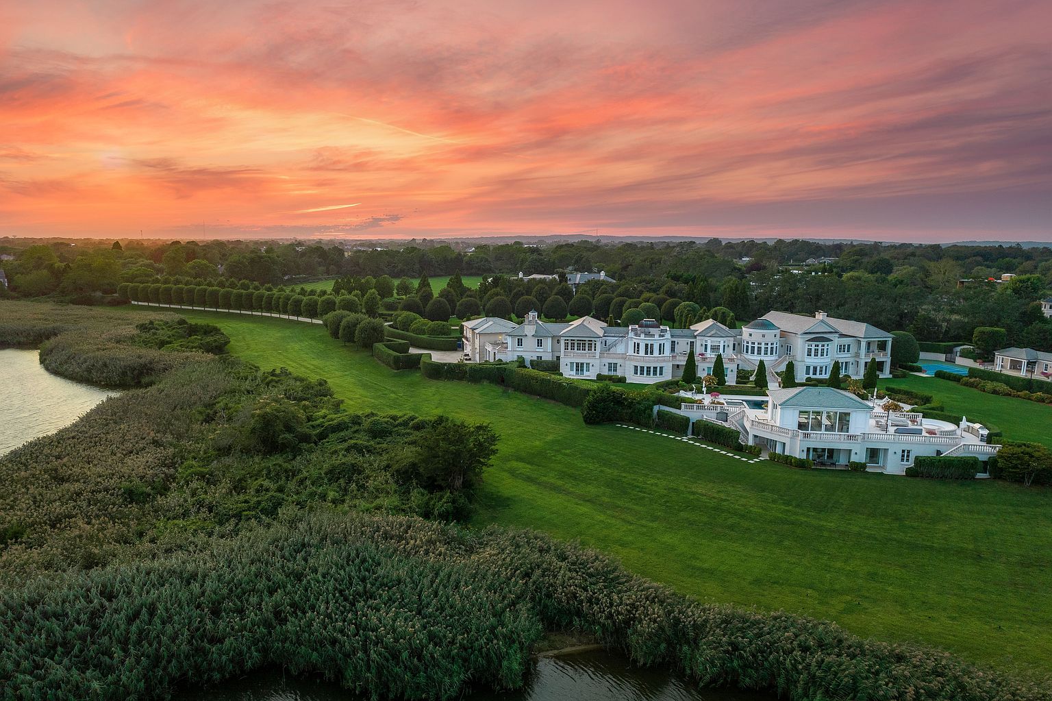 This aerial view showcases a sprawling estate with manicured lawns, lush greenery, and a grand white mansion. The property features a waterfront location with a serene body of water bordering the edge. The sunset sky adds a dramatic and luxurious touch, emphasizing the scale and exclusivity of the property.