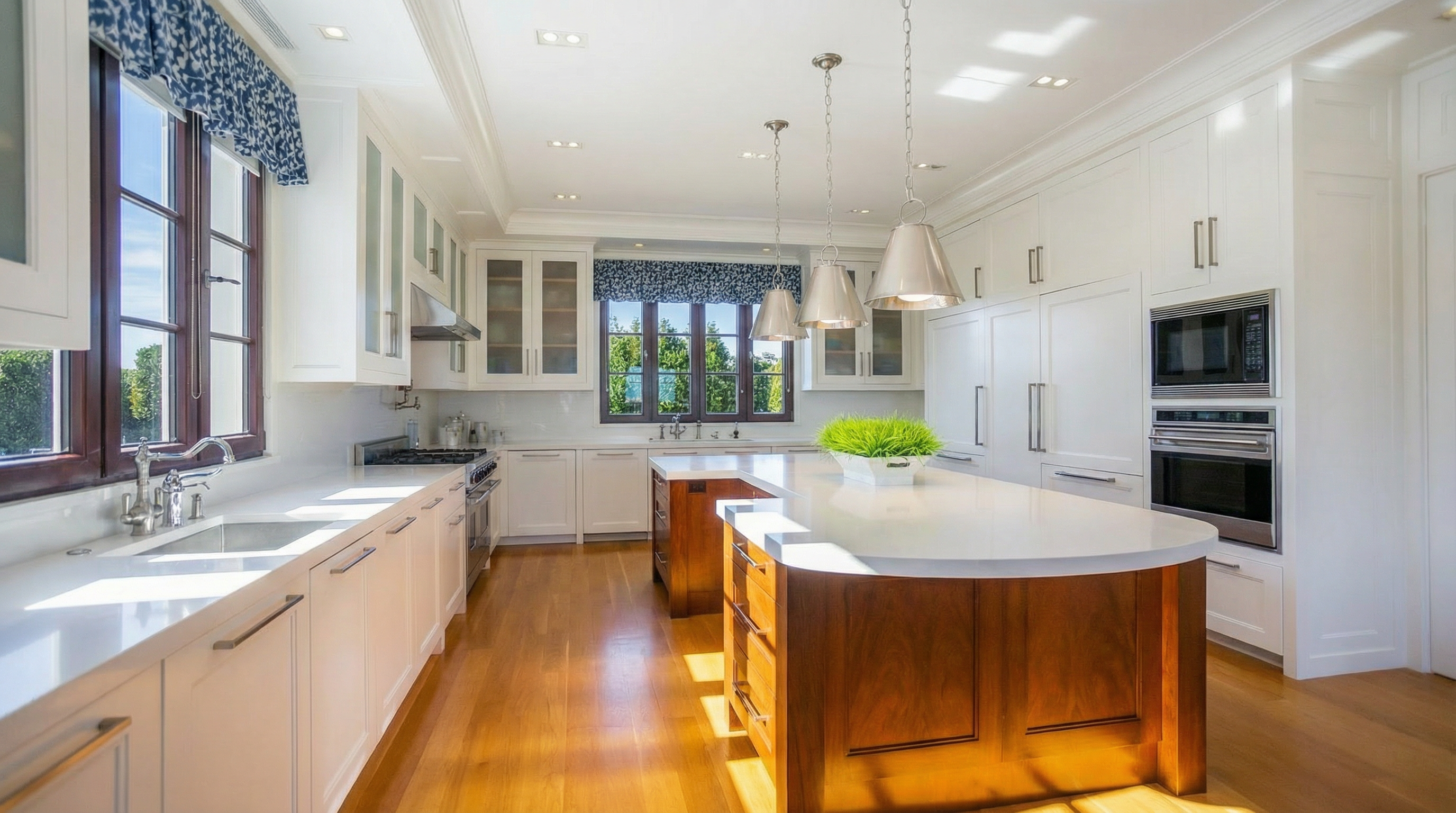 This is a bright and spacious kitchen featuring white cabinetry, stainless steel appliances, and a large wooden island with a white countertop. The kitchen has hardwood floors and ample natural light from the windows, along with modern pendant lighting over the island. It presents a clean and high-end aesthetic typical of a luxury home.
