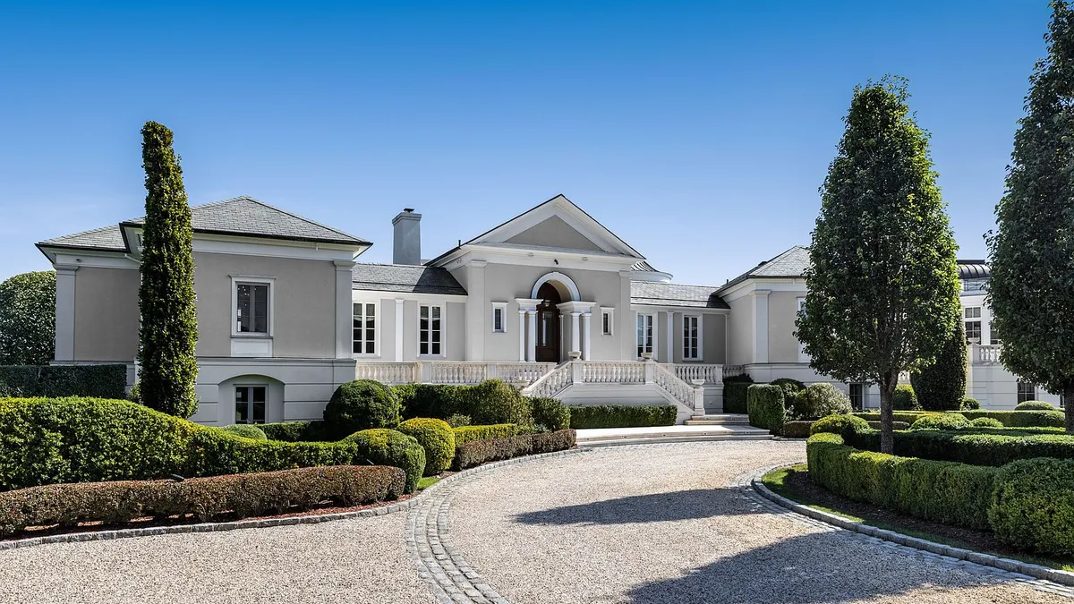 The image features the front exterior of a stately, light-grey colored residence. The architecture includes a symmetrical layout with a grand entrance, multiple windows, and meticulously manicured landscaping with hedges and trees lining the driveway. The property exudes luxury and curb appeal.