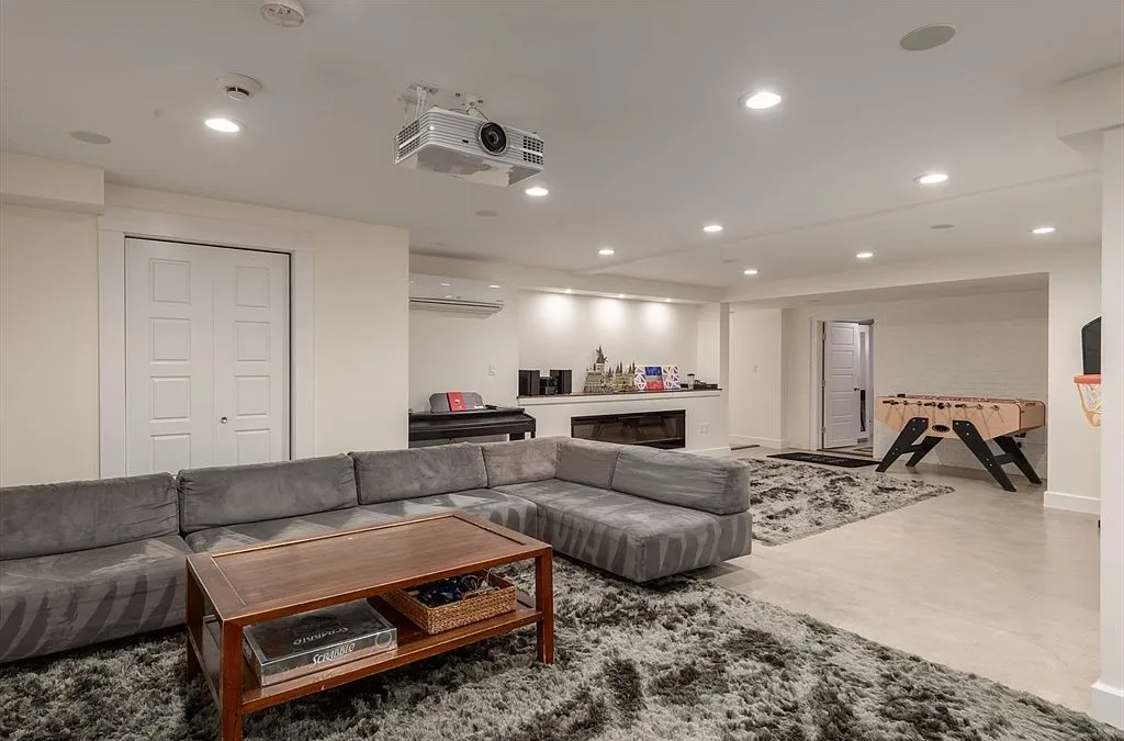 This spacious, finished basement features a large grey sectional sofa, a coffee table with board games, and a plush area rug, making it an ideal recreation or media room. The space is equipped with a ceiling-mounted projector, a foosball table, and built-in shelving near a cozy fireplace. The neutral color palette and recessed lighting create a clean, versatile living area perfect for family activities and guest entertainment.