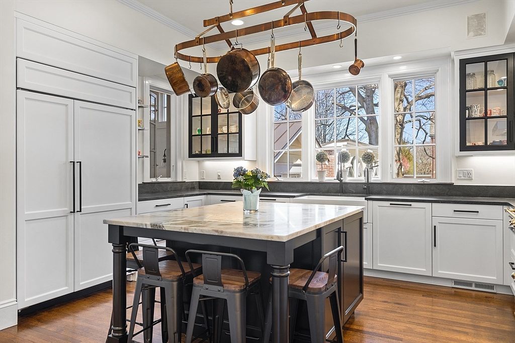 This bright and elegant kitchen features a sophisticated color palette with white perimeter cabinetry accented by a contrasting dark kitchen island topped with a luxurious marble countertop. A statement copper pot rack suspended above the island adds a vintage-inspired charm, while large windows bring in ample natural light. The space is completed by hardwood flooring, sleek metal stools, and integrated high-end appliances, creating a timeless and functional design.