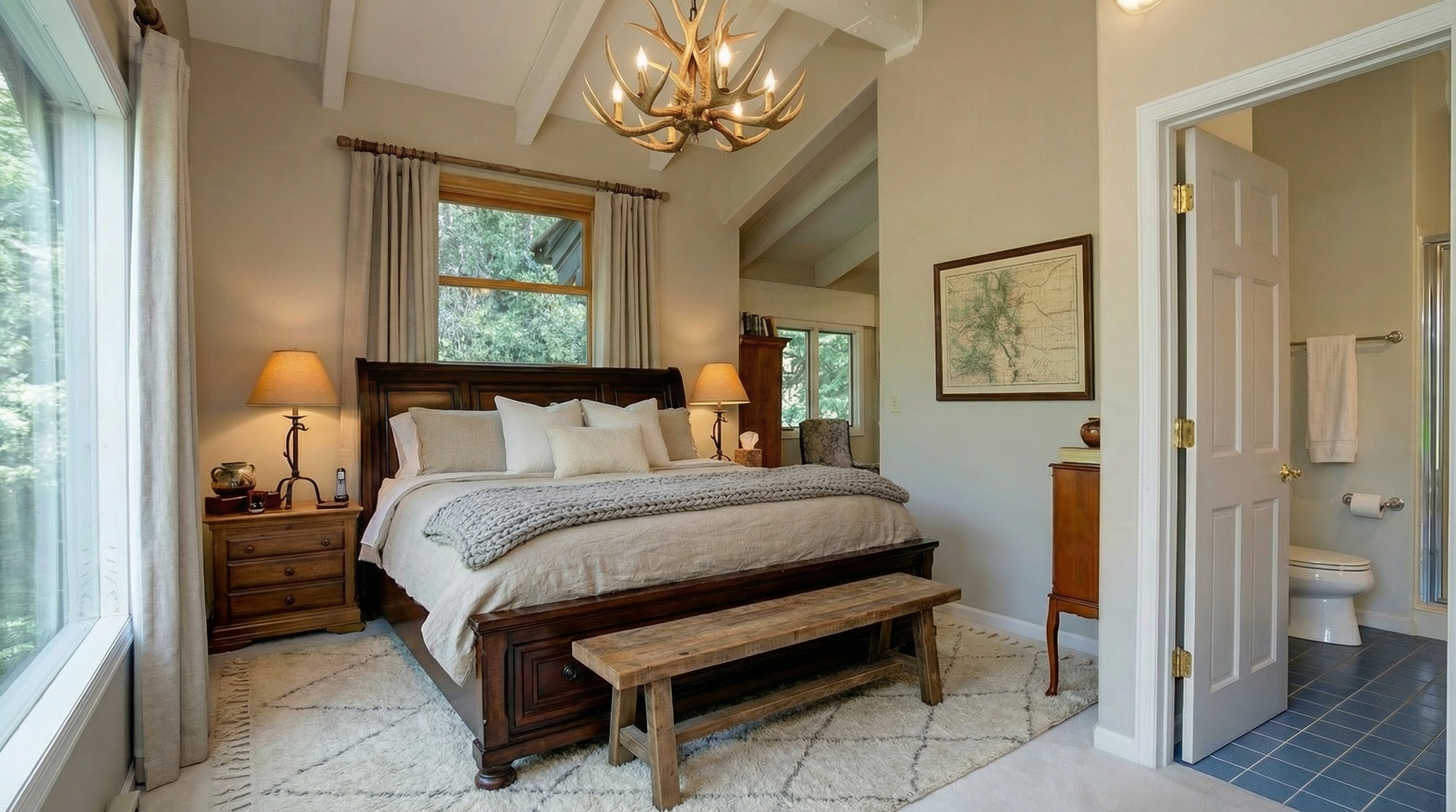 This is a warmly decorated primary bedroom featuring a dark wood bed frame with neutral bedding and a rustic wooden bench at the foot of the bed. The room has a cozy, inviting atmosphere, enhanced by soft lighting from bedside lamps. A hardwood nightstand complements the bedroom's furnishings. There is a view to an en suite bathroom.