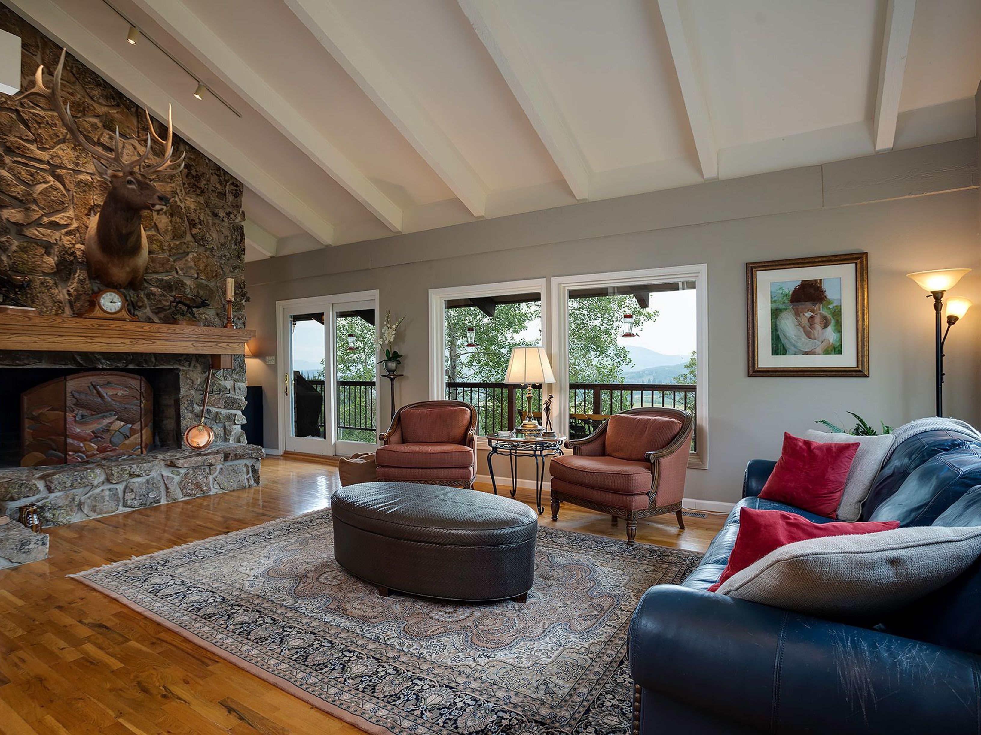 A cozy living room features a stone fireplace with a mounted deer head, a dark leather sofa with red throw pillows, and two armchairs flanking a small table. The room is bathed in natural light from windows overlooking a wooded area, with a neutral-toned area rug adding warmth to the hardwood floors. The beamed ceiling adds an element of architectural interest and height to the space.