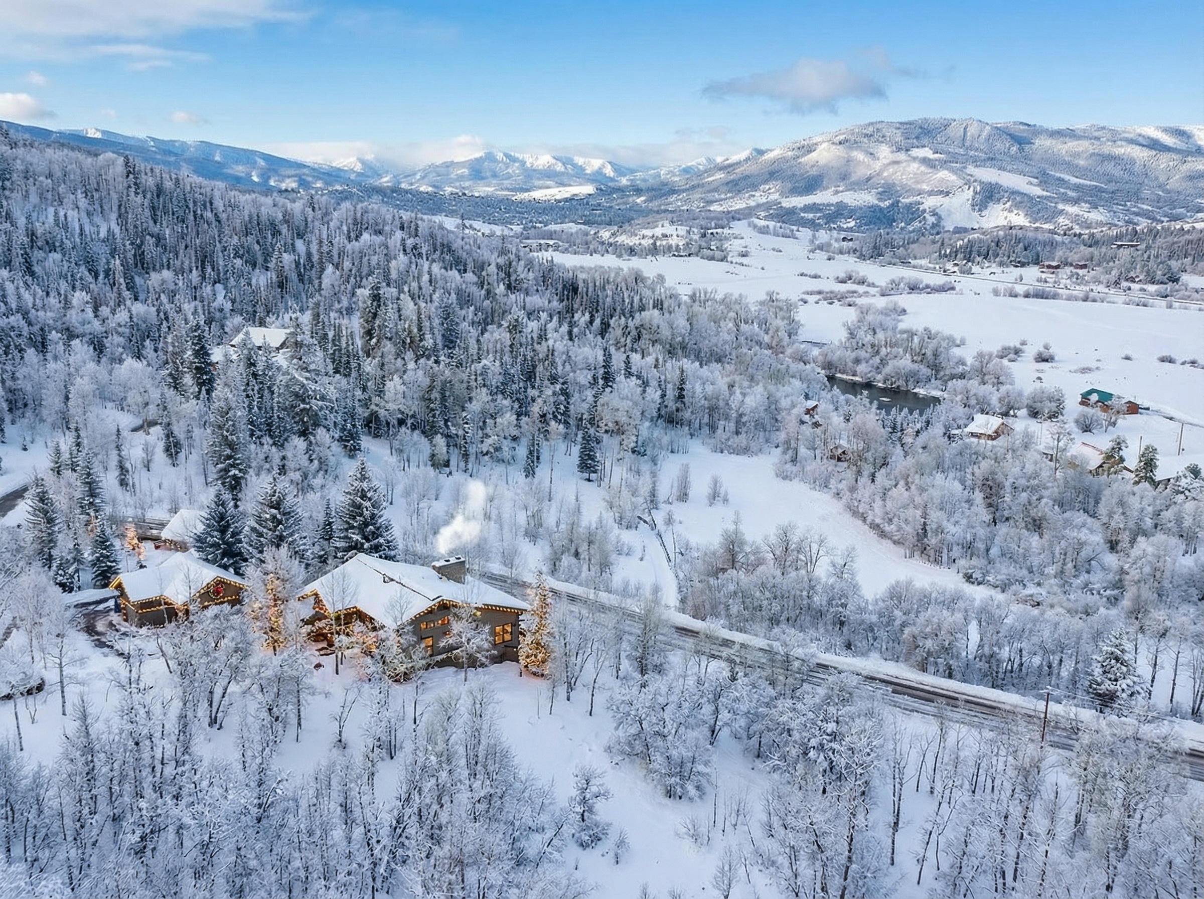 This aerial view showcases a beautiful property covered in snow, giving a winter wonderland impression. The house is adorned with Christmas lights and flanked by snow-covered trees and a sprawling landscape, highlighting the privacy and scenic beauty of the location. The picturesque setting, including distant mountains, emphasizes the property's appeal as a serene escape.