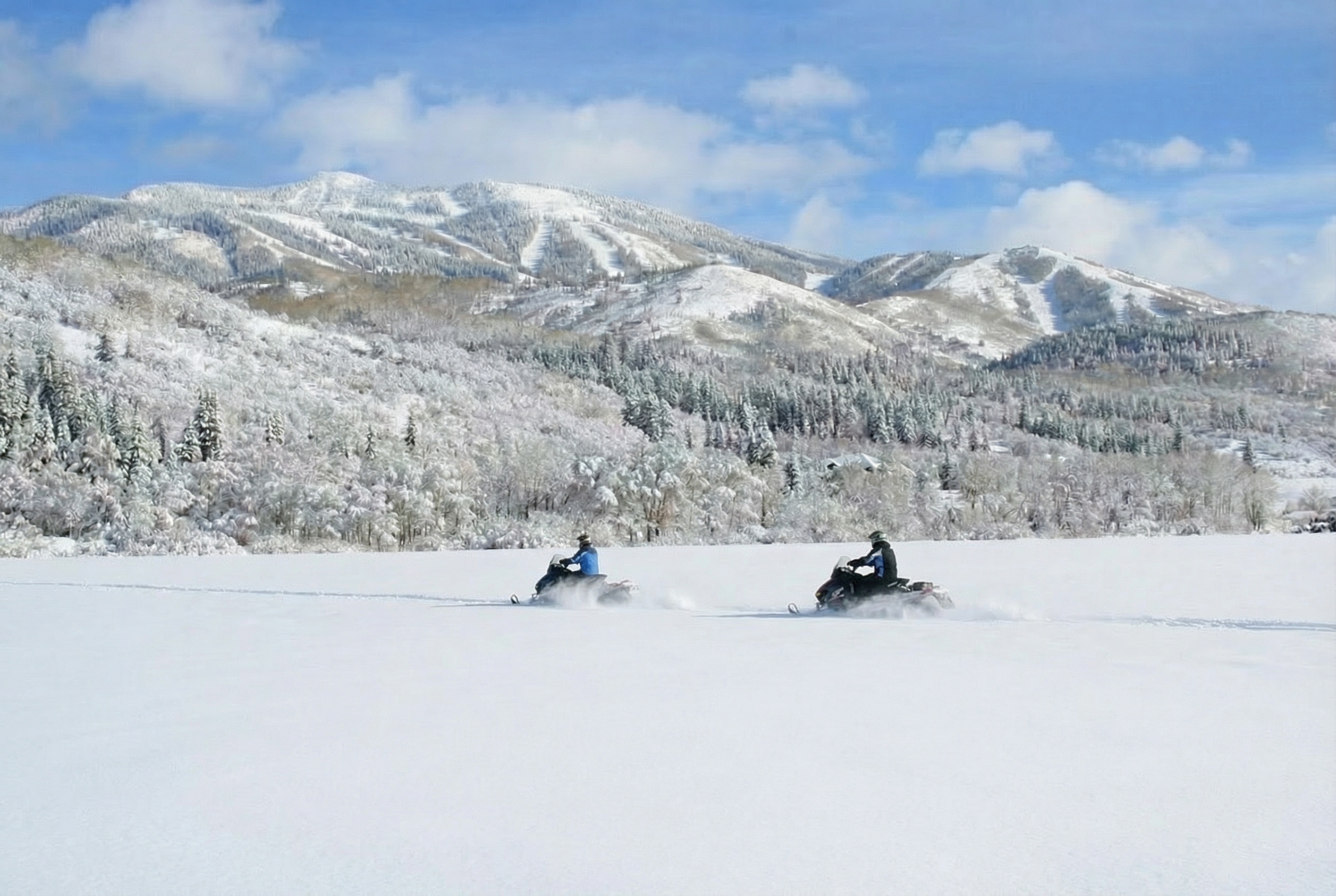 This image showcases a winter landscape featuring snow-covered mountains and trees. Two snowmobiles are traversing the snowy terrain, suggesting recreational opportunities. The scene conveys a sense of peacefulness and outdoor adventure, potentially appealing to buyers interested in winter sports or a tranquil environment.